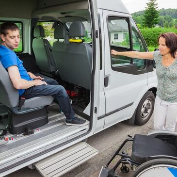 The image depicts a woman assisting a young man, who appears to have mobility challenges, as he sits in a wheelchair accessible vehicle. The woman stands by the open side door of the van, extending her hand towards the man, suggesting she is helping him to step up into the vehicle, which is specifically designed for those with disabilities. The van features ramps for easy access, highlighting its suitability for individuals requiring mobility assistance. The surrounding area indicates a peaceful suburban environment, potentially in or near Moncrieff, where His Grace Care operates, further emphasising the supportive and community-focused nature of their home assistance services. This visual aligns with the mission of His Grace Care, showcasing their commitment to providing compassionate, accessible support to those in need, ensuring comfortable transportation and assistance for their clients.