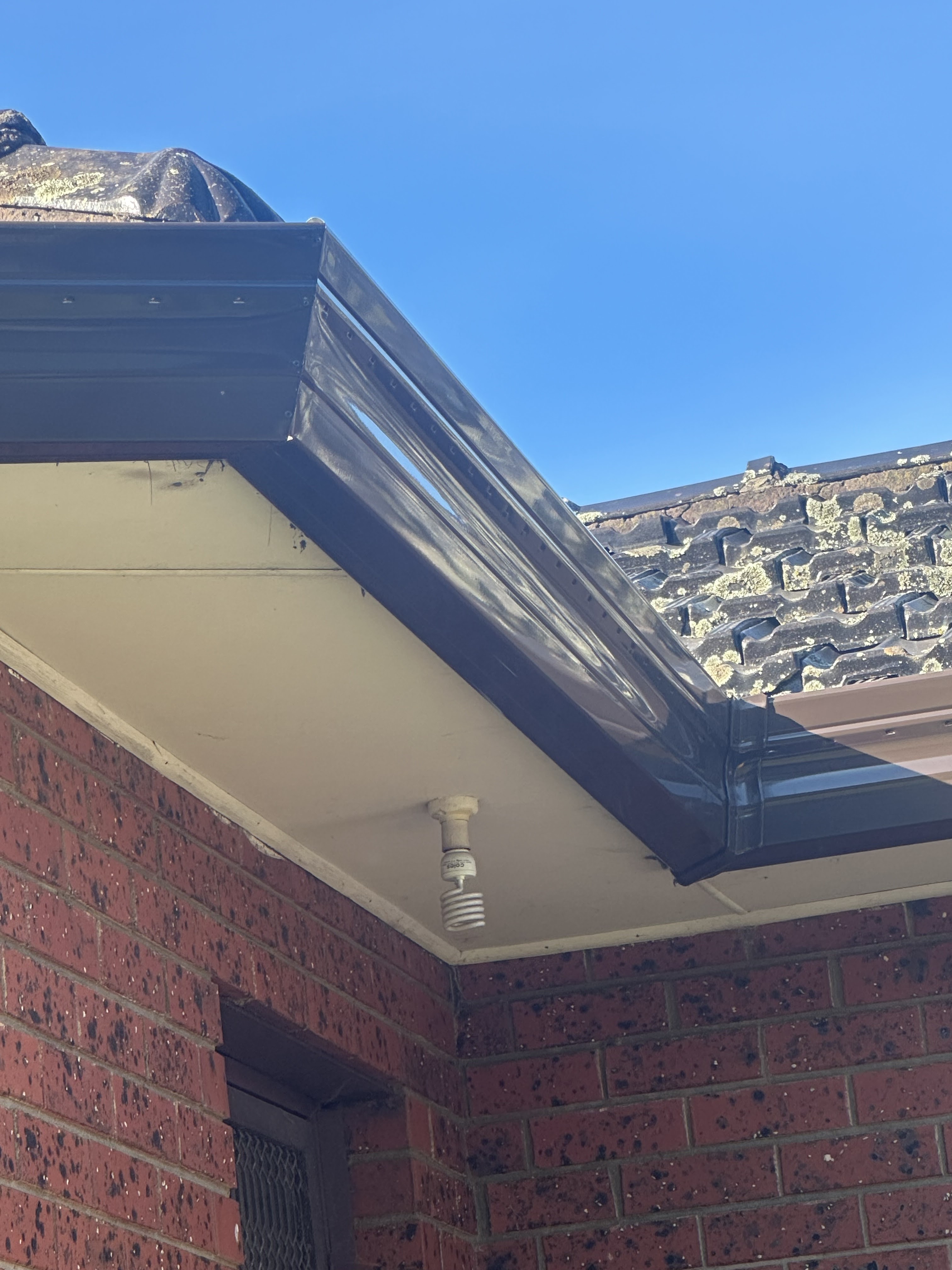 The image showcases a close-up view of the eaves of a building, likely taken during a roofing inspection or maintenance check. The soffit, with a seamless edge, is painted in a light colour and appears to be in good condition, indicating recent work or upkeep. The guttering, which is glossy and black, suggests a focus on aesthetic appeal as well as functionality, effectively directing rainwater away from the structure. A hanging light fixture is visible, hinting at practical lighting solutions in the area. The blue sky in the background provides a bright backdrop, perfect for highlighting the quality roofing services provided by DJL Roofing and Gutters, indicating a well-maintained environment that exemplifies their commitment to excellence in roofing and gutter systems.