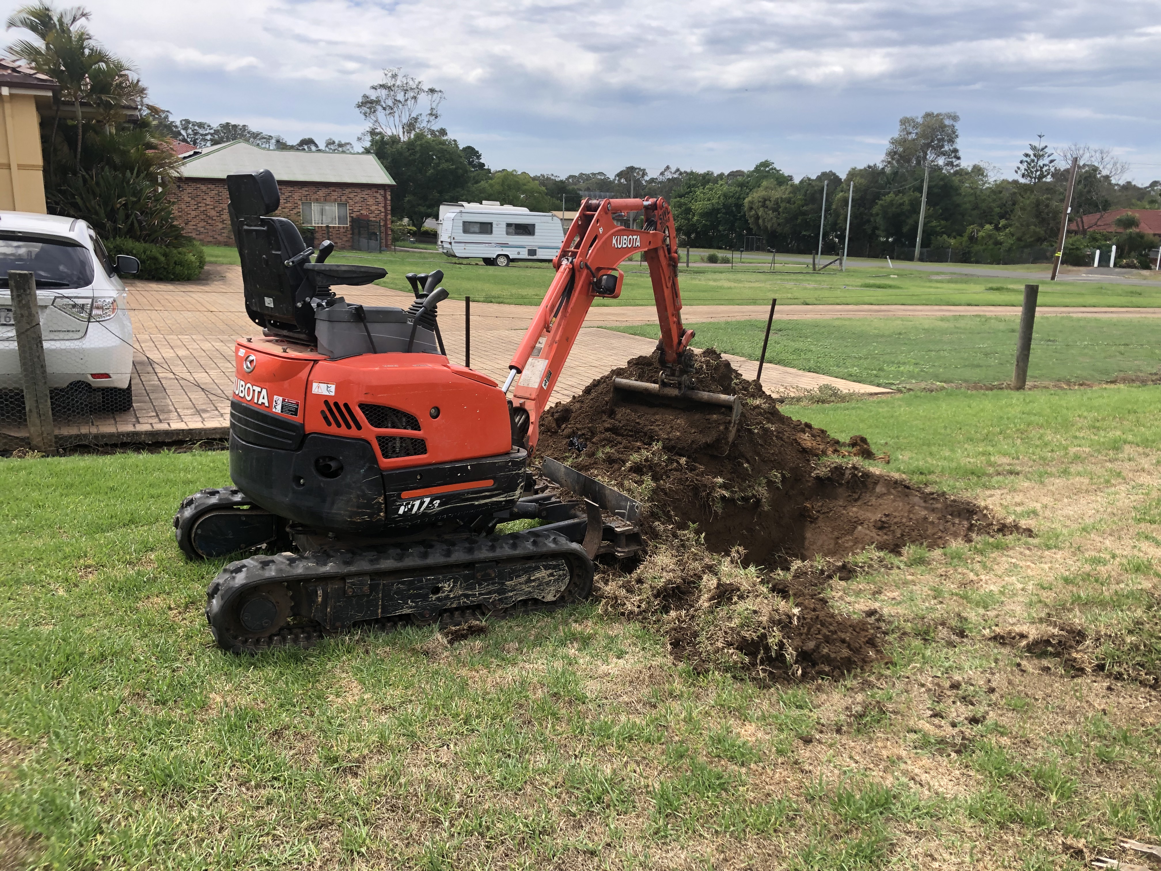 The image features a compact excavator with a digging arm, parked on a grassy area with a mound of freshly excavated soil beside it. In the background, there are residential properties. This setup suggests work related to plumbing, possibly for laying pipes or installing a hot water system, typical of services such as those provided by Hot Water Services Penrith, especially in areas like Mulgoa and surrounding suburbs.