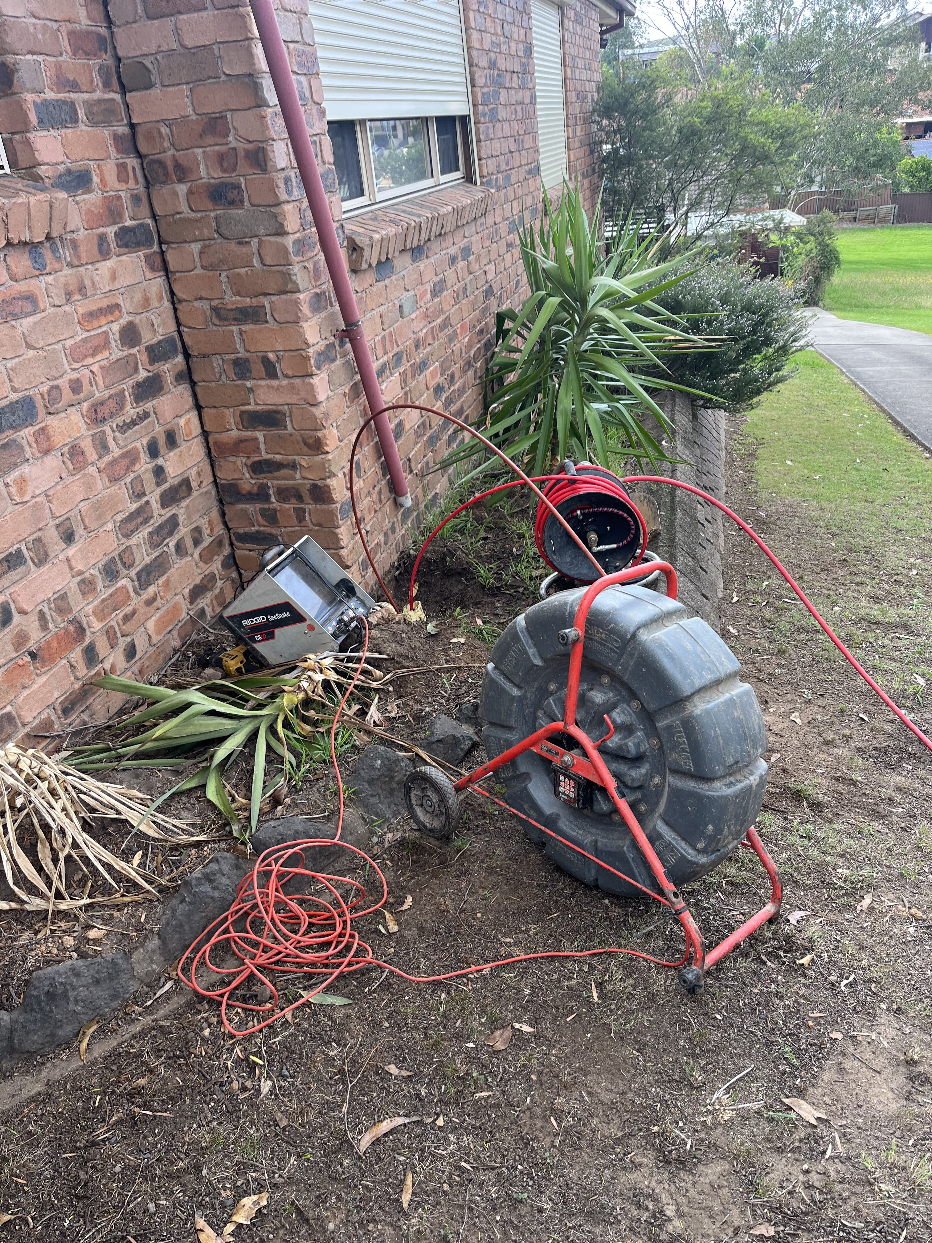 The image displays plumbing equipment typically used for drain cleaning and maintenance. In the foreground, there is a large, wheeled drain cleaning machine with a coiled hose, likely intended for unclogging pipes. The machine is connected to a power source, indicated by the surrounding electrical cords. Nearby, some landscape elements such as a plant and dirt suggest an outdoor setting, possibly near the property's foundation. The scene is indicative of plumbing work, where tools are prepared for a job, likely involving clearing a blockage in a residential drainage system.