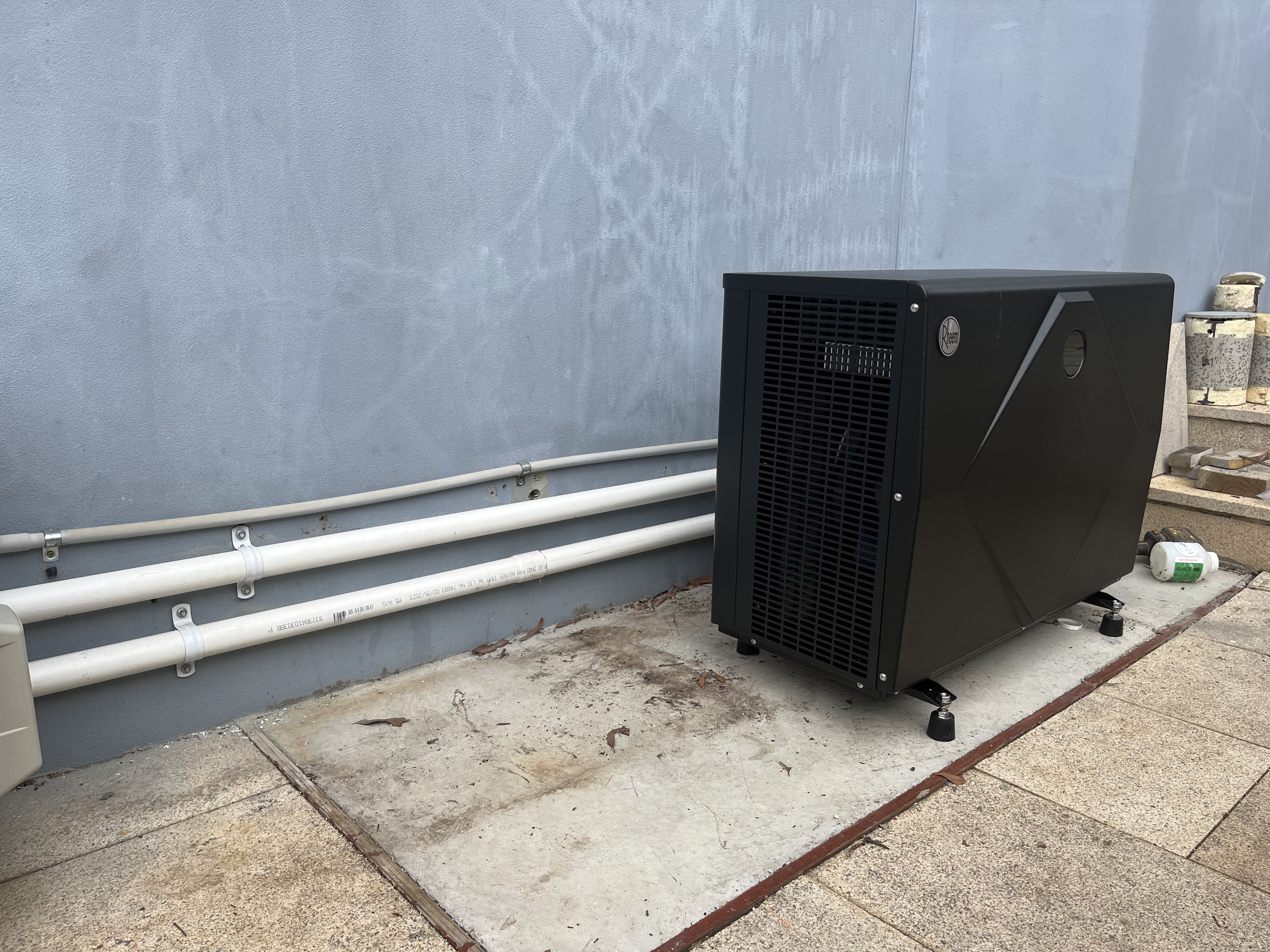 The image depicts an outdoor installation of a large, black heat pump or hot water system, situated against a plain wall. The unit is robust and appears to be designed for efficient energy use, likely for heating water in residential settings. Two white pipelines extend along the wall, which may connect the unit to the water supply or drainage system. The surrounding area consists of concrete paving, indicating a stable installation surface, and there are remnants of construction materials scattered around, possibly from recent plumbing work. This setup aligns with the services provided by Hot Water Services Penrith, indicating their expertise in installing and maintaining hot water systems in residential areas.