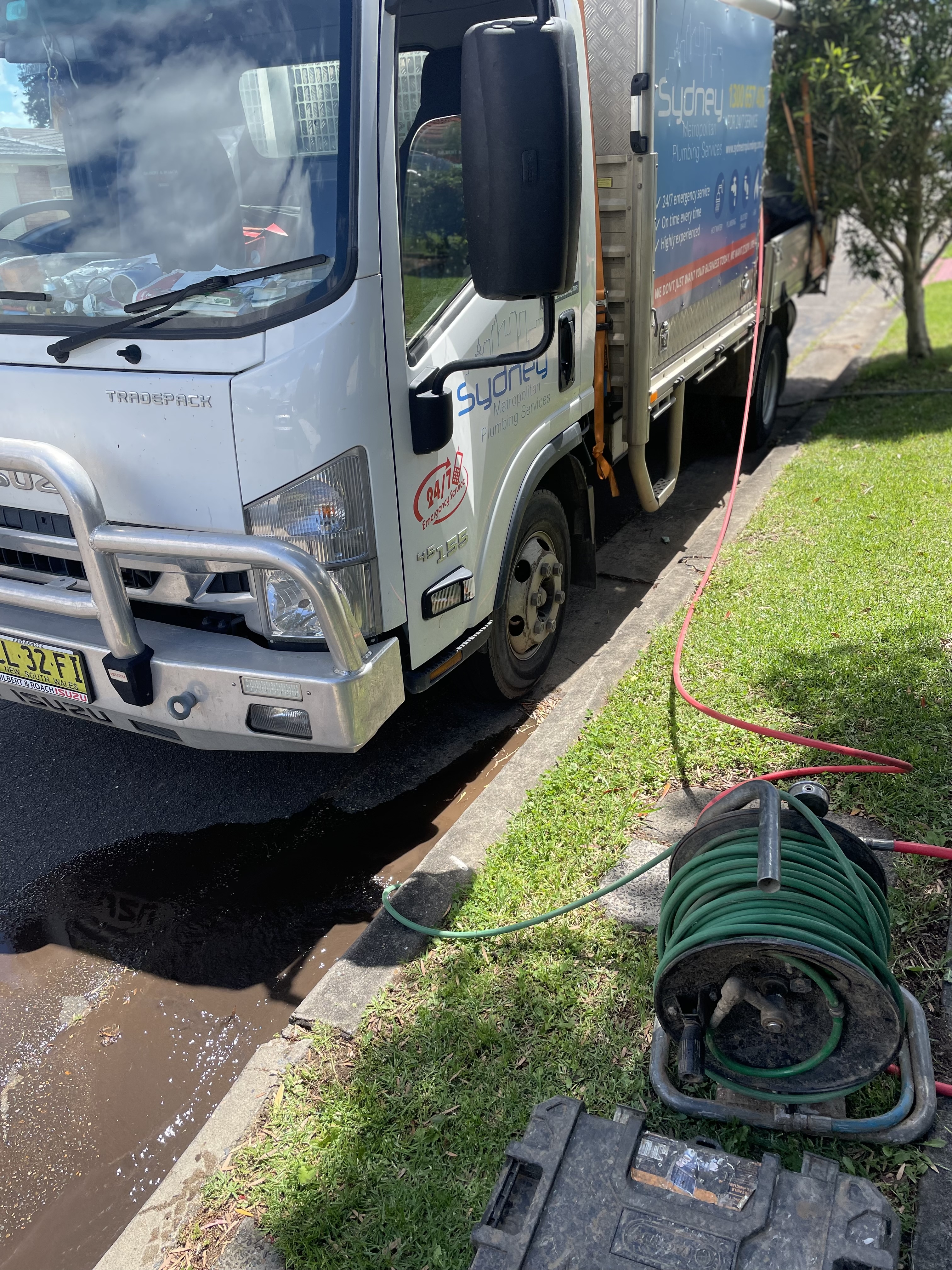 The image shows a plumbing truck parked on the street, with a hose connected to the vehicle. The truck is labelled with 'Sydney Plumbing', indicating its plumbing services. A hose reel is positioned on the ground, suggesting ongoing plumbing work, likely related to drainage or hot water services in the nearby area. The truck appears to be ready for service, highlighting the company's active role in providing plumbing solutions.