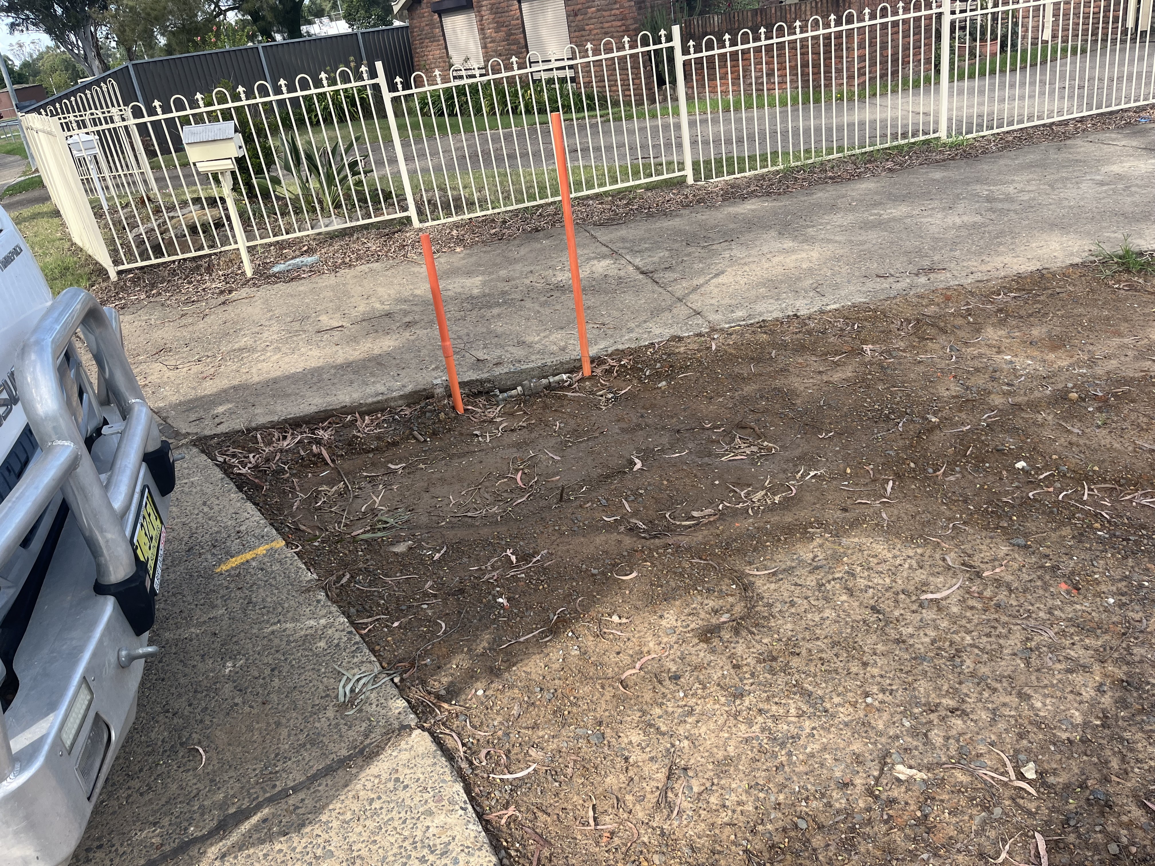 The image shows a section of pavement or a driveway with two orange markers standing vertically in the ground. The area appears to be cleared of debris and is mostly bare earth, suggesting preparation work for plumbing or landscaping. The surroundings include a white vehicle visible on the left side, and in the background, there is a residential property and a white fence. This setup could indicate the location for upcoming plumbing services or installations, likely related to hot water systems, as this business operates in that domain.