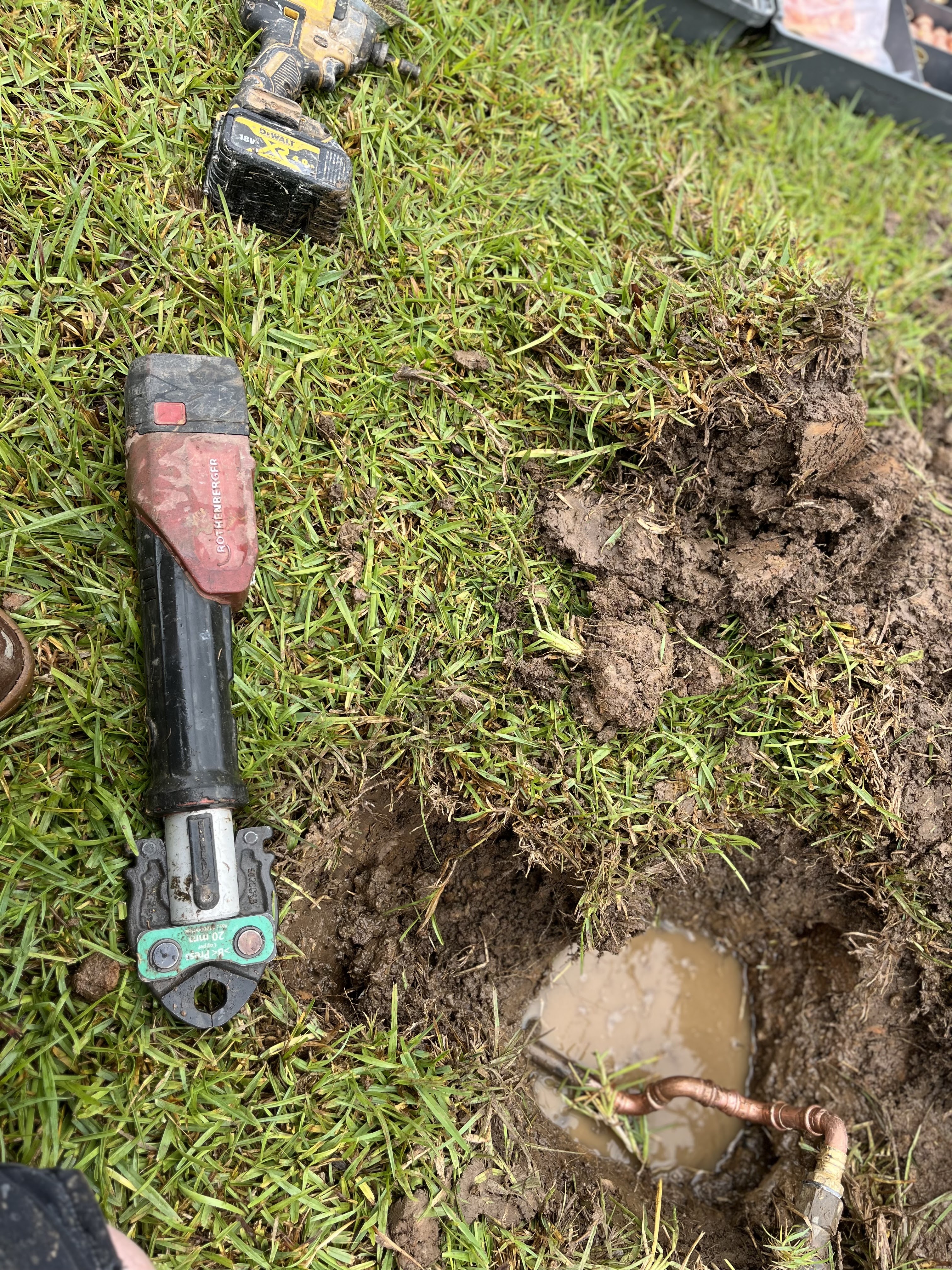 The image shows a trench or hole in a grassy area with water pooling at the bottom. Next to the hole lies a plumbing tool, likely a pipe cutter or similar device, indicating maintenance or repair work on underground water lines. There appears to be a copper pipe partially exposed in the mud, suggesting a plumbing issue that may involve repairing or replacing this pipe. The surrounding environment is typical of a suburban area, possibly indicating that the plumbing work is related to a residential property.