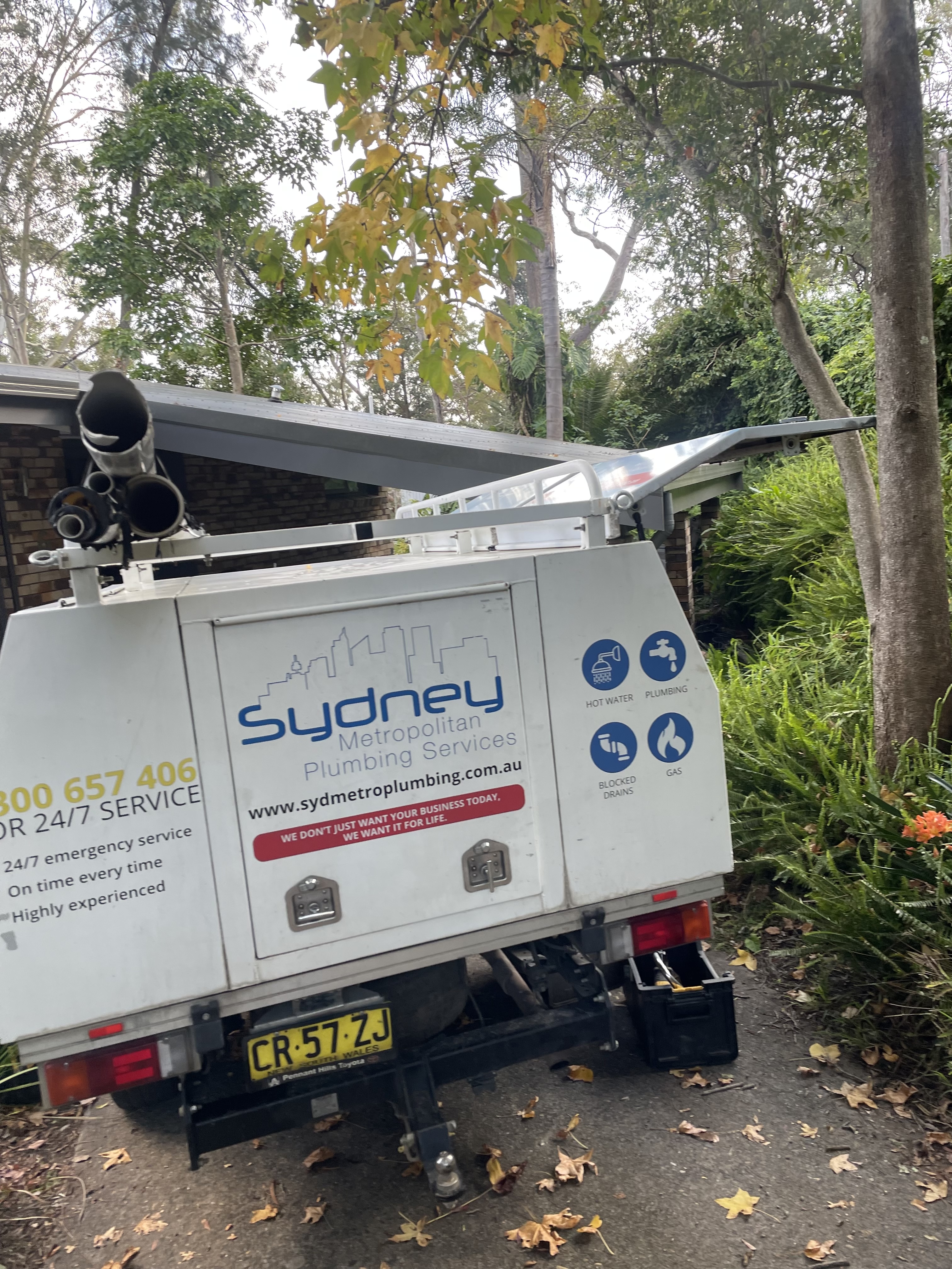 The image showcases a plumbing service vehicle parked in a bushland setting. The rear of the van features a logo for 'Sydney Metropolitan Plumbing Services', indicating it's likely used for plumbing-related tasks. The vehicle has several pipes visible on the roof, suggesting it is equipped for plumbing jobs, perhaps related to hot water systems. The background features lush greenery, typical of suburban landscapes in Mulgoa, NSW.