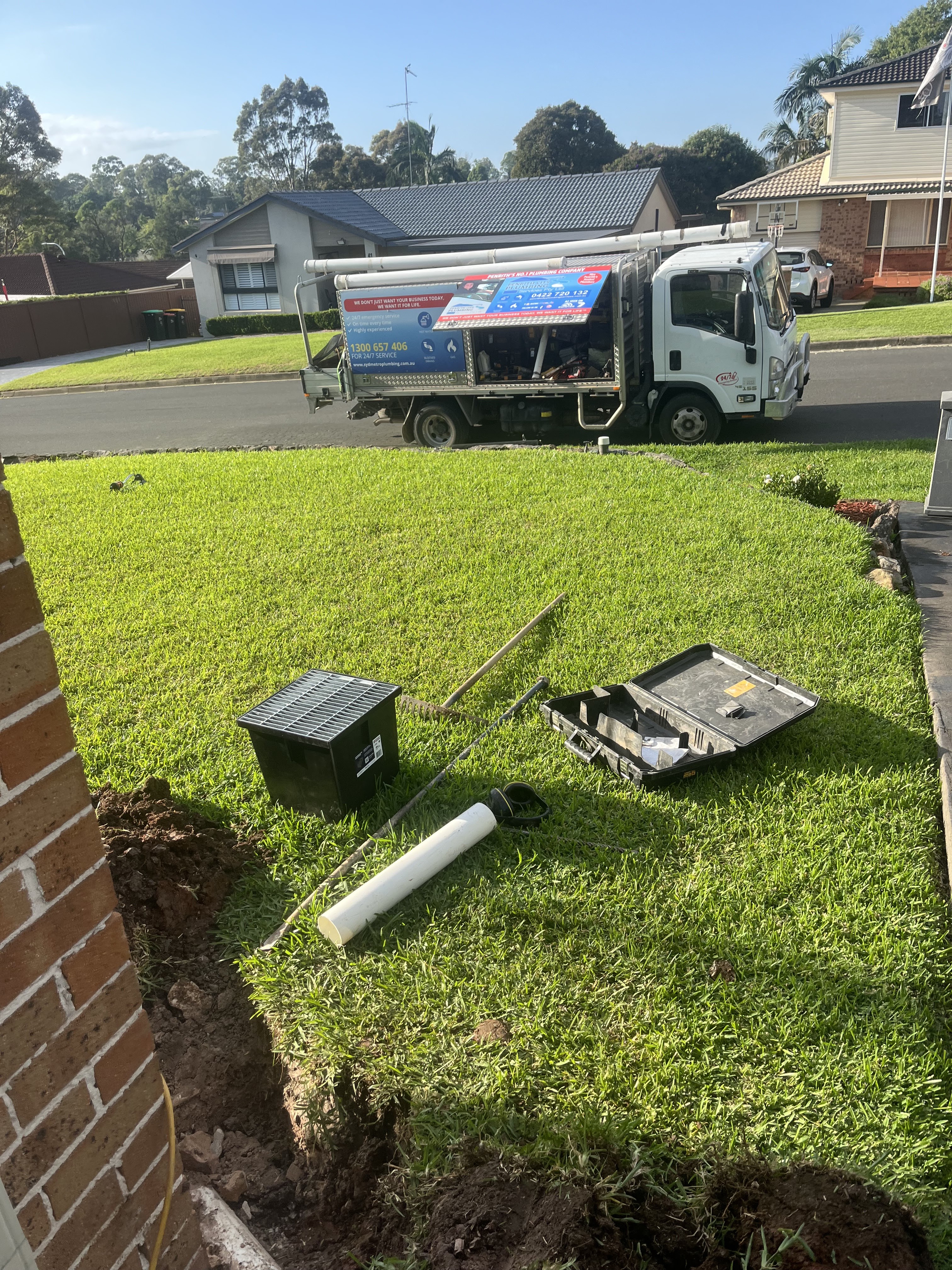 The image shows a plumbing service vehicle parked on a residential street in Mulgoa. The truck features branding that indicates it's associated with Hot Water Services Penrith, highlighting their plumbing expertise. Nearby, there is a construction scenario: tools and equipment are scattered on the grass, including a black toolbox, a white plastic pipe, and a digging implement. The ground appears disturbed, suggesting ongoing plumbing work. The surroundings are suburban, with houses visible in the background, hinting at a community-focused service.
