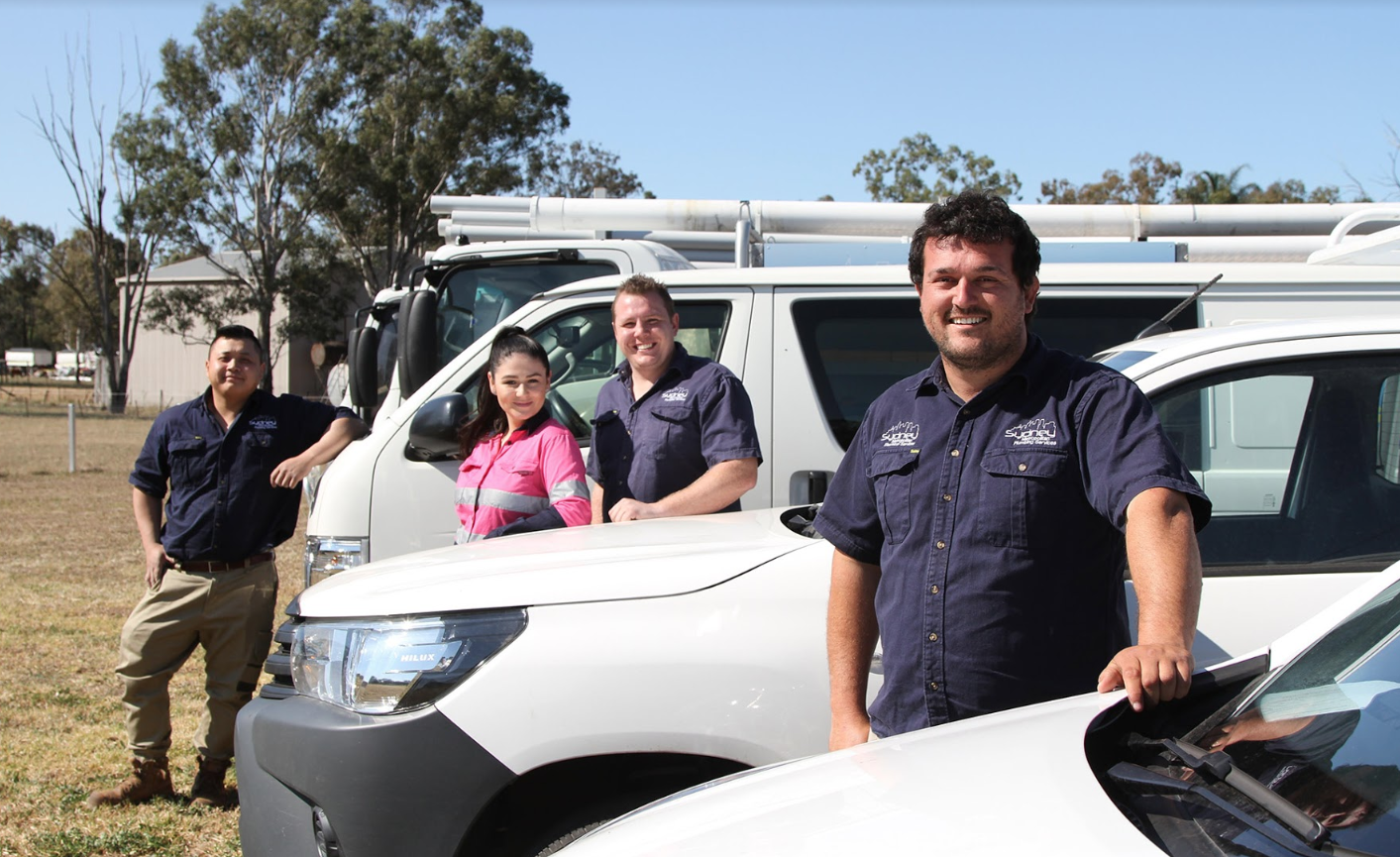 The image features four team members of a plumbing business, standing confidently by their work vehicles in an outdoor setting. The individuals are dressed in matching blue company shirts, presenting a professional appearance. The background shows greenery, suggesting a rural or semi-urban environment. The vehicles, prominently displayed, reflect the plumbing service's operational focus, likely used for various plumbing tasks in the vicinity. The team's camaraderie and readiness to assist customers are evident in their expressions and posture, enhancing the business's image as reliable and approachable.