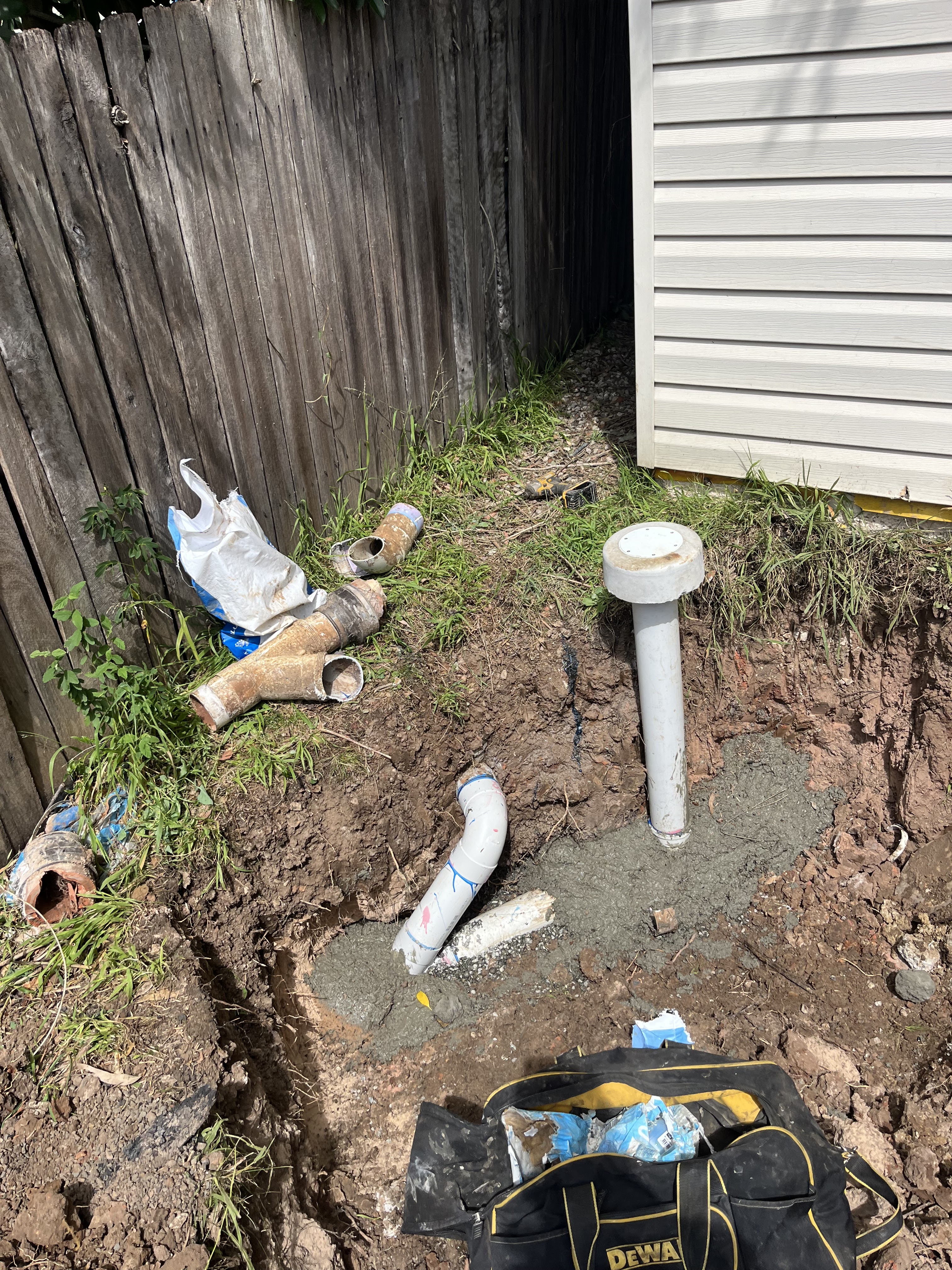 The image depicts an excavation site with exposed plumbing pipes, indicating ongoing plumbing work. There are several discarded pipe fragments scattered around the area, highlighting recent repair or installation activities. A new white vertical pipe appears to have been installed, set in a bed of concrete for stability, suggesting it might be a part of drainage or water supply system. The surrounding area features overgrown grass and remnants of debris, which may require cleanup after the plumbing work is completed. Items related to the plumbing job, like a black tool bag, are visible, indicating that professional plumbing tools and equipment are being used.
