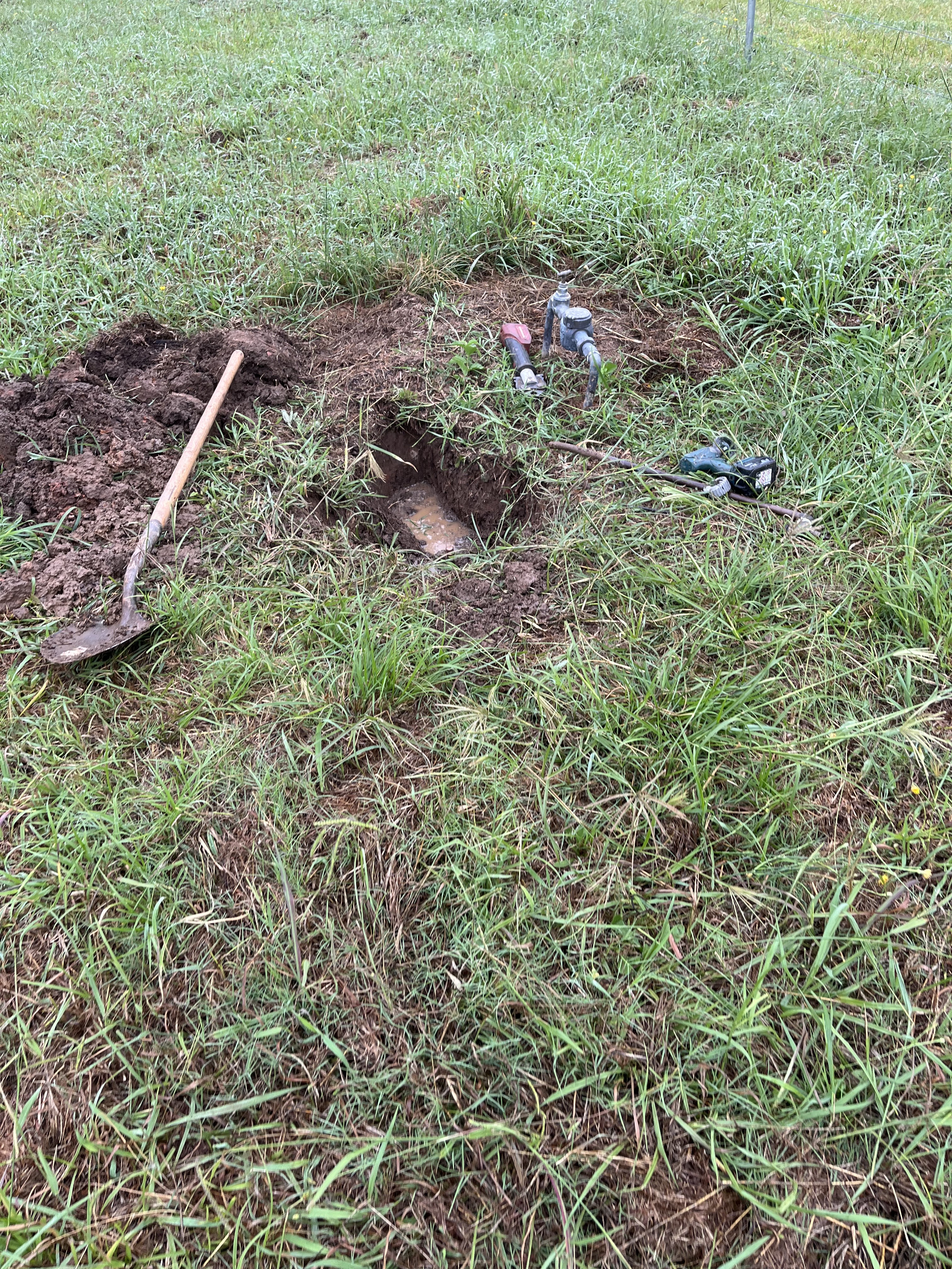 The image depicts a section of land where work is being carried out for plumbing purposes. A hole has been dug into the ground, revealing some pipes possibly related to drainage or water supply. Nearby, there are tools including a shovel and other plumbing tools, indicating an ongoing plumbing repair or installation process. The surrounding area is grassy, suggesting a residential or garden setting.
