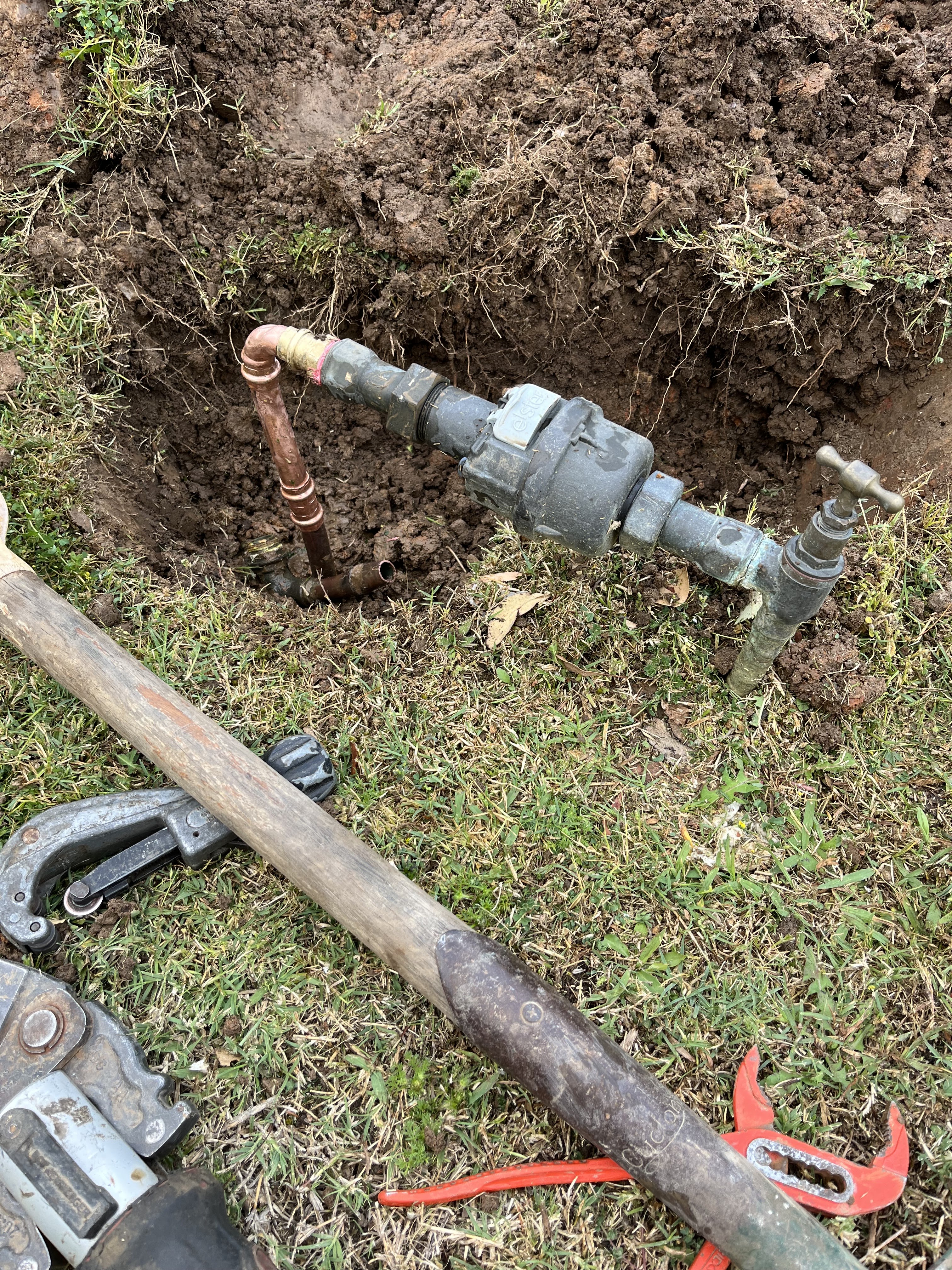 The image shows an underground plumbing setup with exposed pipes and fittings. There is a copper pipe connected to a valve, and several tools, including a wrench, are visible nearby. The area around the pipes is dug out, indicating ongoing work, possibly for maintenance or repairs on the water supply system. This is typical for a professional plumber addressing plumbing issues, ensuring proper functionality.