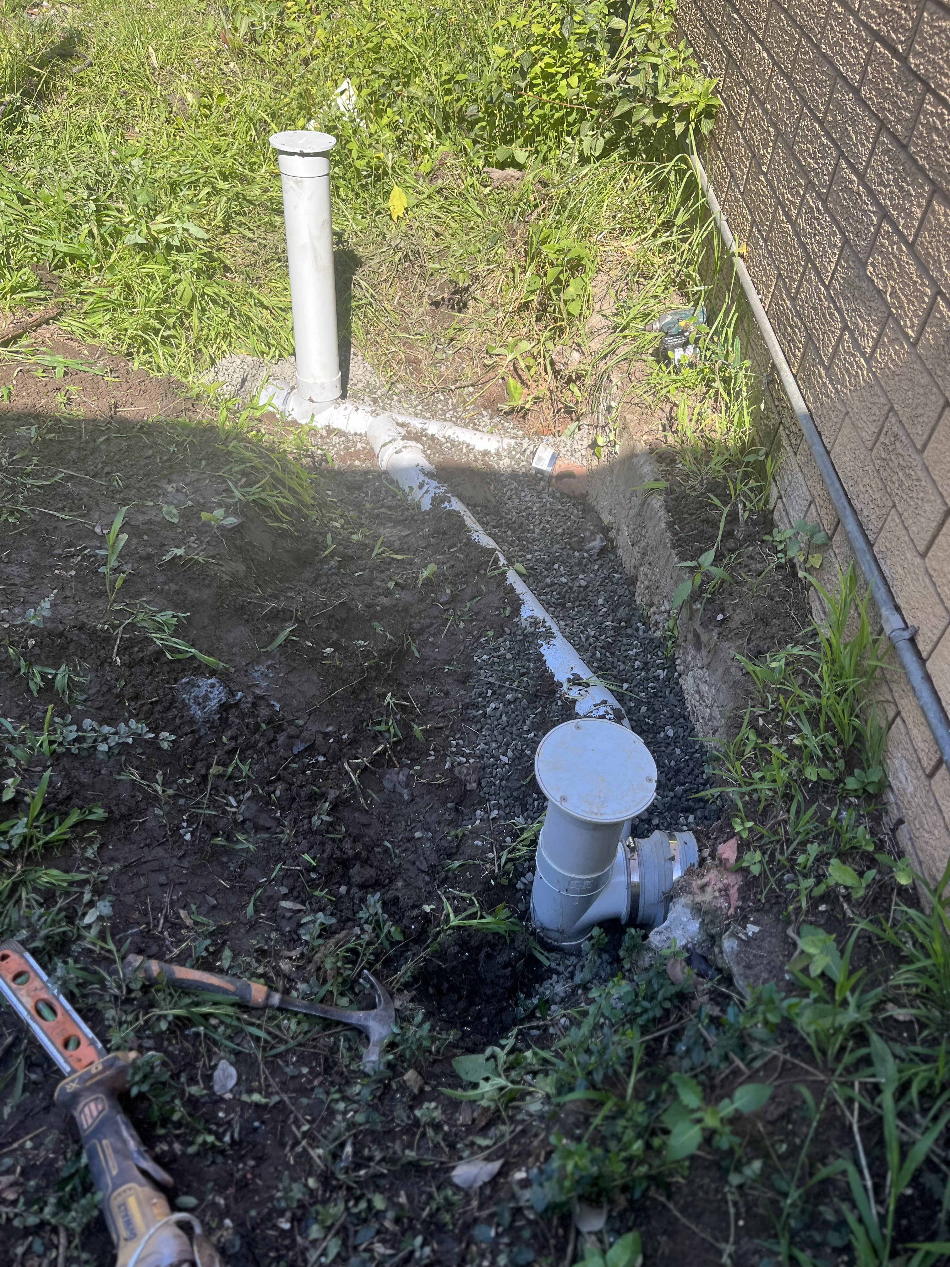 The image shows a partially excavated area in a garden or yard where plumbing work is being conducted. Visible are grey PVC piping systems set into the ground, likely part of a drainage or sewer system. The pipes are connected at a junction, indicating a functional drainage setup. Surrounding the pipes is disturbed soil, with grass and weeds present, suggesting recent digging. This scene conveys the ongoing work of a plumbing service, specifically focused on drainage solutions.