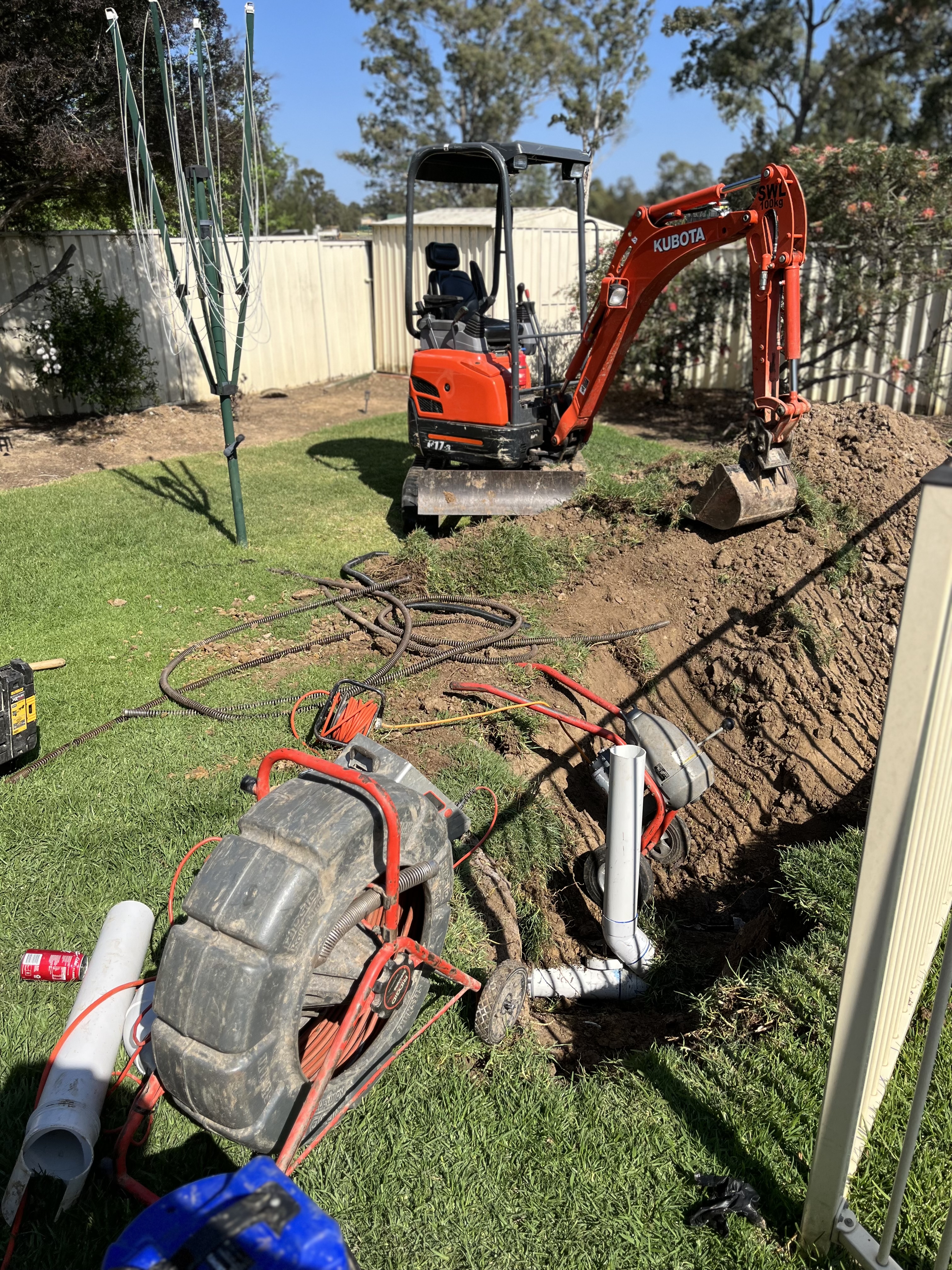 The image showcases a small construction site in a residential backyard where a mini excavator is parked. The machine is primarily used for digging, likely in preparation for plumbing work under the lawn. Surrounding the excavator are various tools and pipework including a large wheel, hoses, and sections of PVC pipe that indicate ongoing drain clearing and plumbing activities. The area appears well-maintained, with green grass and a wooden fence in the background, suggesting a suburban environment. This setting reflects the type of outdoor work typically undertaken by plumbing services in the region.