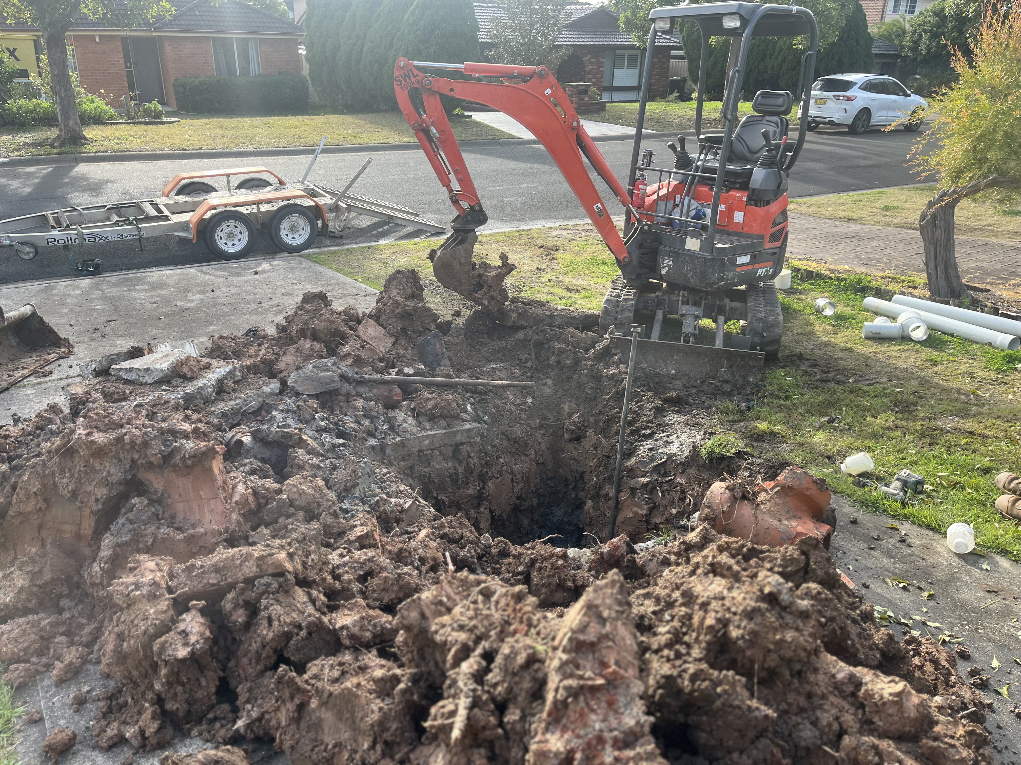 The image depicts a heavy machinery excavator digging into a large, muddy hole in a residential area, likely to access or repair underground pipes. The excavator is parked on a driveway, and there's debris scattered around, indicating ongoing plumbing work. In the background, there's a trailer and some additional plumbing supplies. This scene is typical for plumbing services that require extensive digging, particularly for drainage issues.