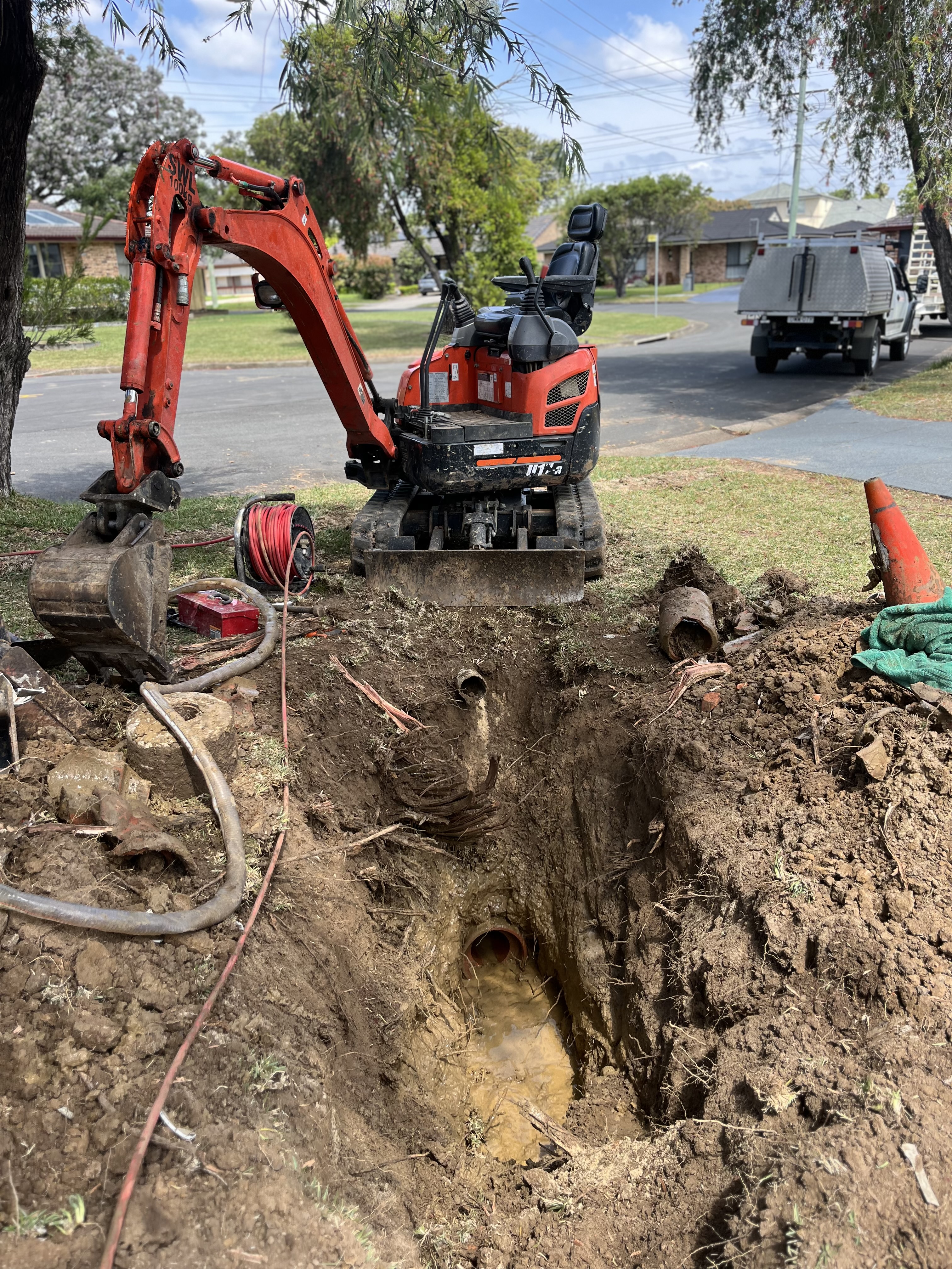 The image showcases a small excavator parked over a dug trench, indicative of ongoing plumbing or drainage work. The trench appears to be deep, revealing pipes that might require attention, possibly for clearing blockages or performing repairs. Nearby tools and equipment suggest that a thorough drainage problem is being addressed. The setting is residential, with a clear view of the street and other properties in the background.