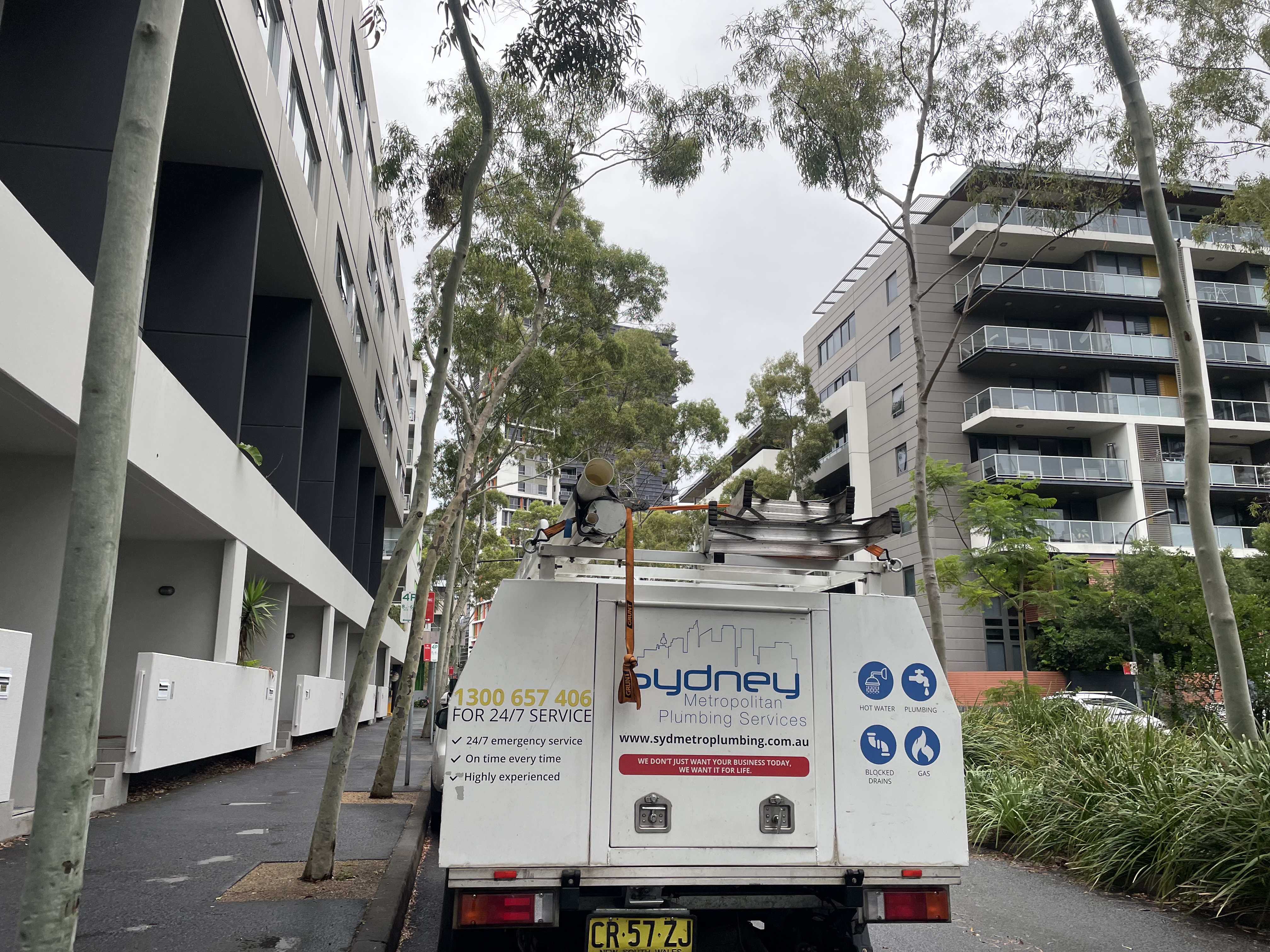 The image depicts a plumbing service truck from a company named 'Sydney Metropolitan Plumbing Services'. The vehicle is parked in a residential area, surrounded by modern apartment buildings and tall trees. The truck is equipped for plumbing tasks, possibly featuring tools or equipment on its roof. The weather appears to be overcast, suggesting it may be raining.