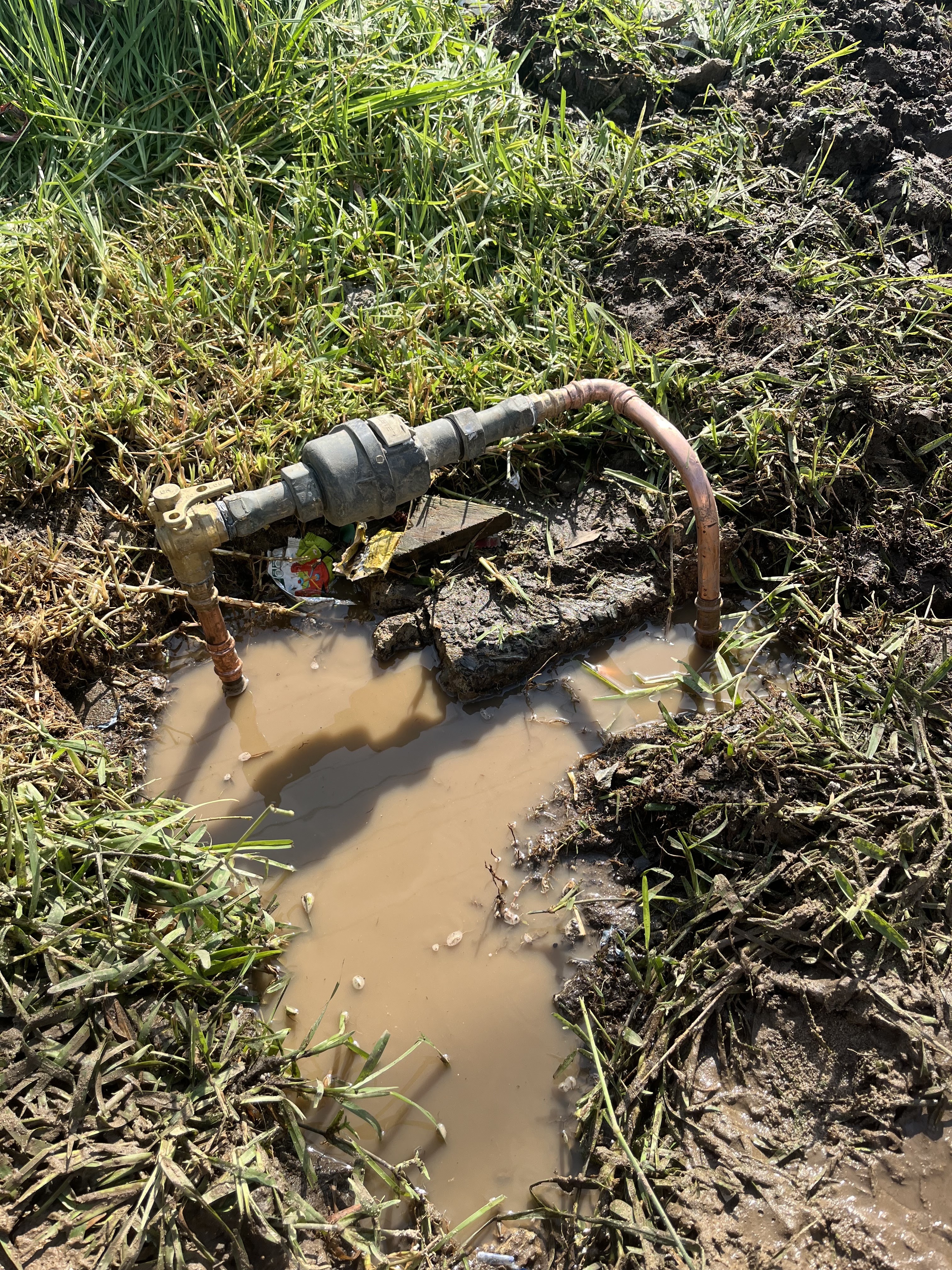The image depicts a damaged plumbing fixture situated in an excavation site, surrounded by muddy grass. Water is pooling in the dug-out area, indicating a possible leak or break in the piping system. The visible copper pipes suggest that this may be part of a water supply line. The unkempt terrain further hints at emergency plumbing work, possibly required due to flooding or a significant water issue.