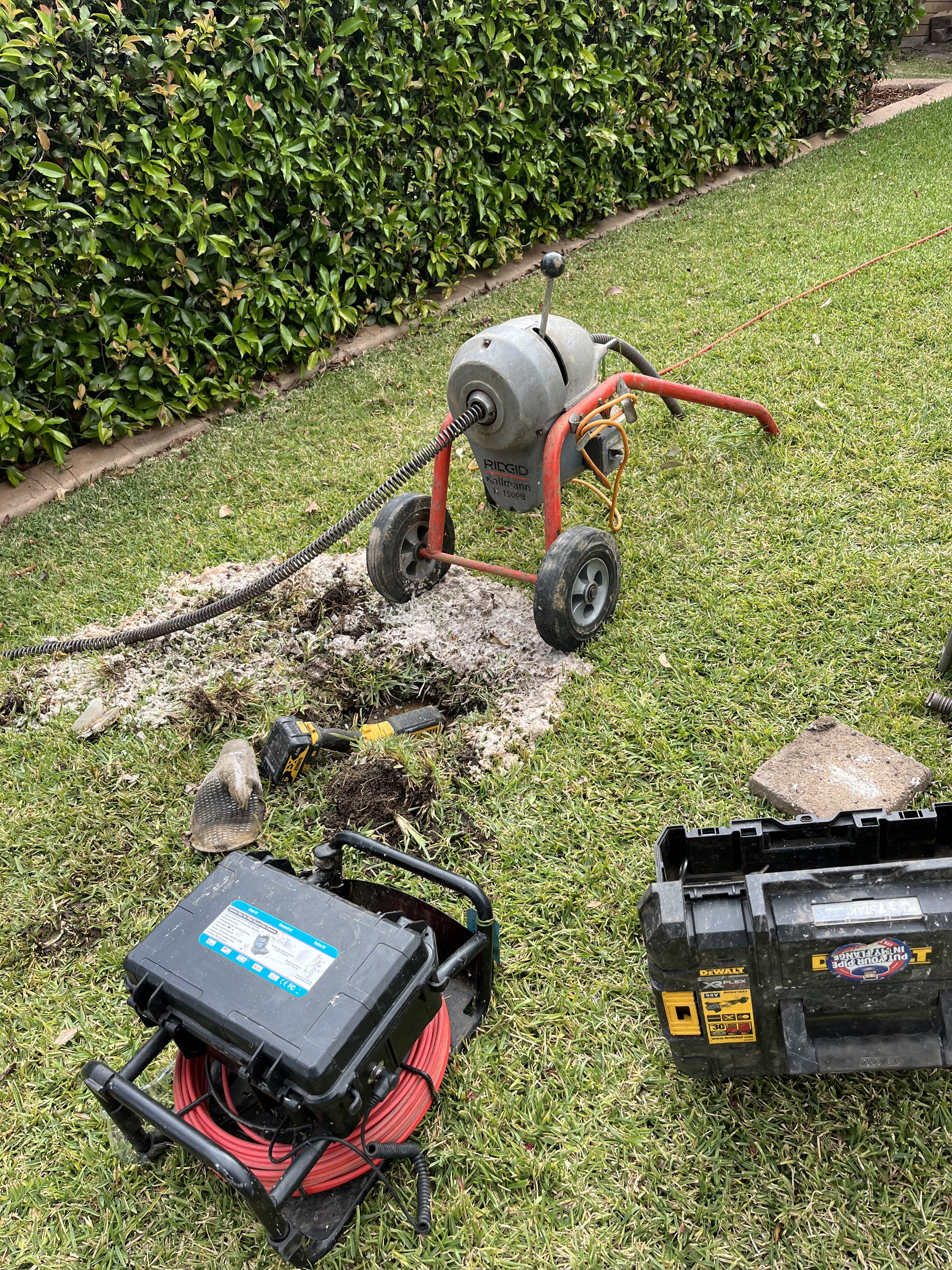 The image depicts a scene in a garden where a drain clearing setup is in progress. A drain clearing machine is positioned on a small patch of dug-up grass, indicating ongoing maintenance or repair work. The machine is connected to a flexible auger or cable, likely used for clearing blockages in drains. Scattered nearby are tools essential for plumbing tasks, including a tool box and additional equipment such as a reel of cable. The surrounding area is neatly kept, filled with green grass and hedges, typical of residential landscaping in Mulgoa.