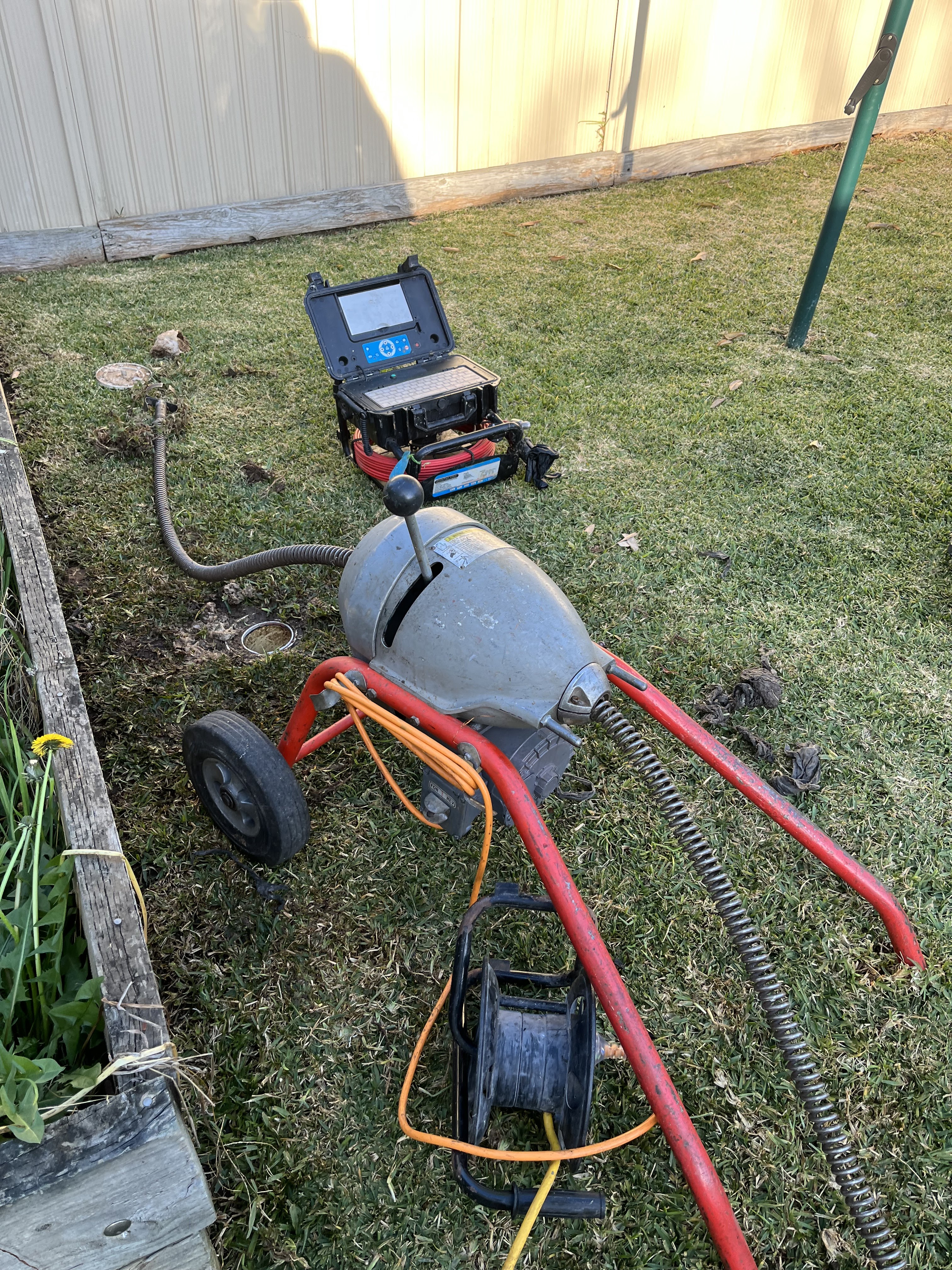 The image shows a drain clearing machine set up in a grassy area, likely outside a property. The machine, which has a distinctive long cable and reel, is used for clearing blocked drains or pipes. Nearby, a portable power source is positioned, suggesting it is being powered for operation. The environment appears to be well-kept, which may indicate a residential property receiving plumbing services. The setting is practical for the services provided by Drain Clearing Penrith, highlighting their expertise in resolving plumbing issues in outdoor areas.