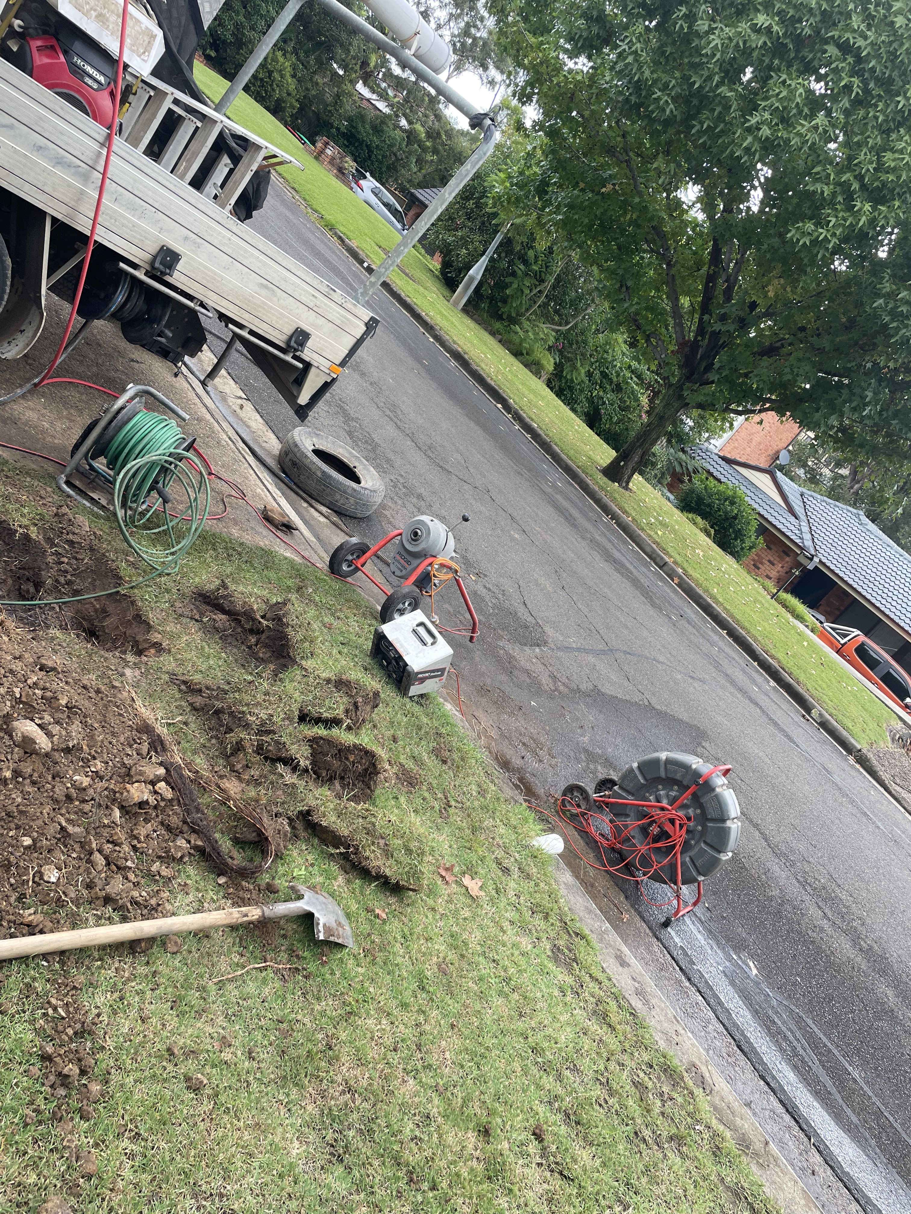 The image shows a plumbing job site on a suburban street. There are several tools and equipment related to drain clearing, including a drain snake, coils of piping, and a portable generator. The ground is disturbed, indicating recent digging, likely to access or clear a blocked drain. A truck is parked nearby, possibly containing additional tools and supplies. The area appears well-maintained with trees lining the street, typical of the Mulgoa and Penrith suburbs.