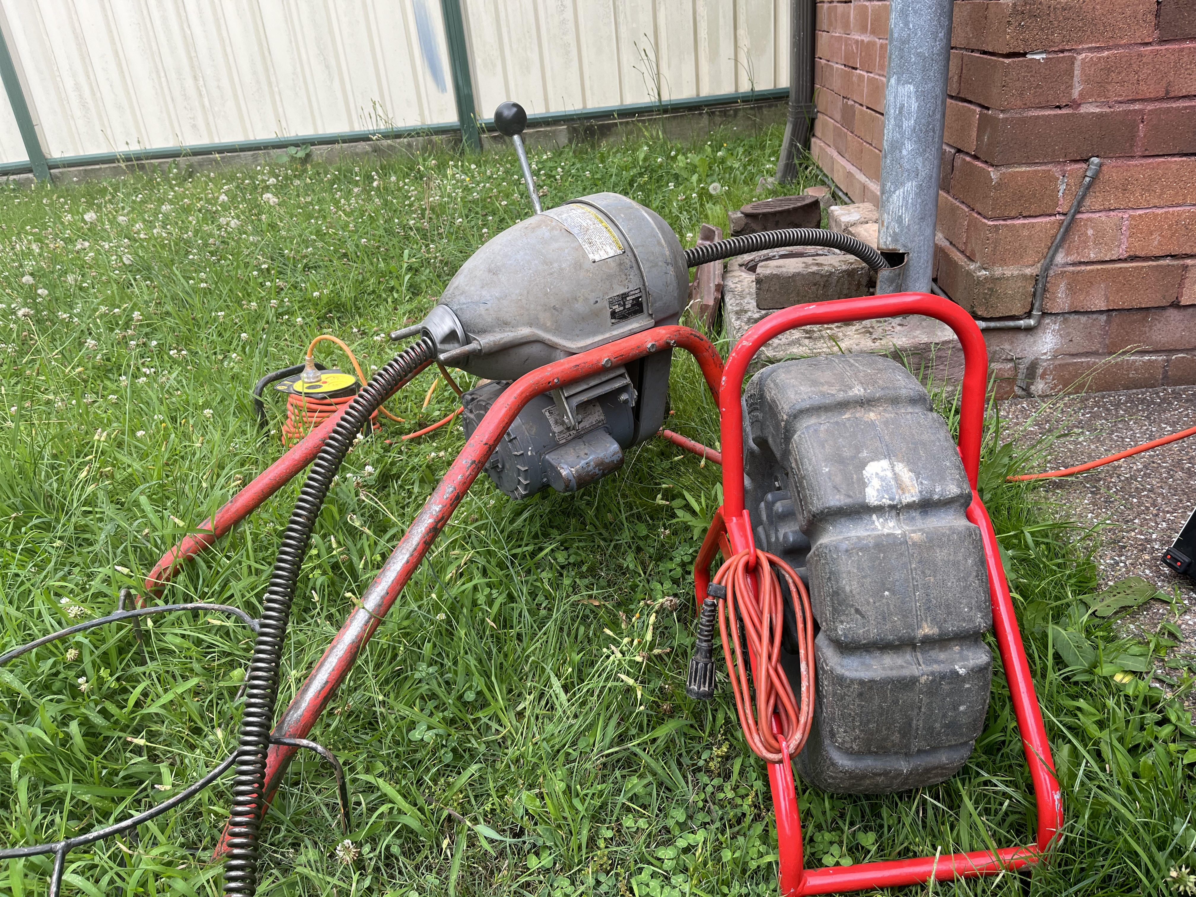 The image showcases a drain cleaning machine, specifically a cable auger, with a prominent red frame. It appears to be placed on grassy ground, indicating it is likely used for outdoor plumbing tasks. The equipment includes an electric motor, a rotating cable, and a coiled hose meant for clearing blocked drains, showcasing the tools of a plumbing business like Drain Clearing Penrith. This type of equipment is essential for effectively unclogging sewer lines and drain systems, especially in residential areas like Mulgoa and Penrith.