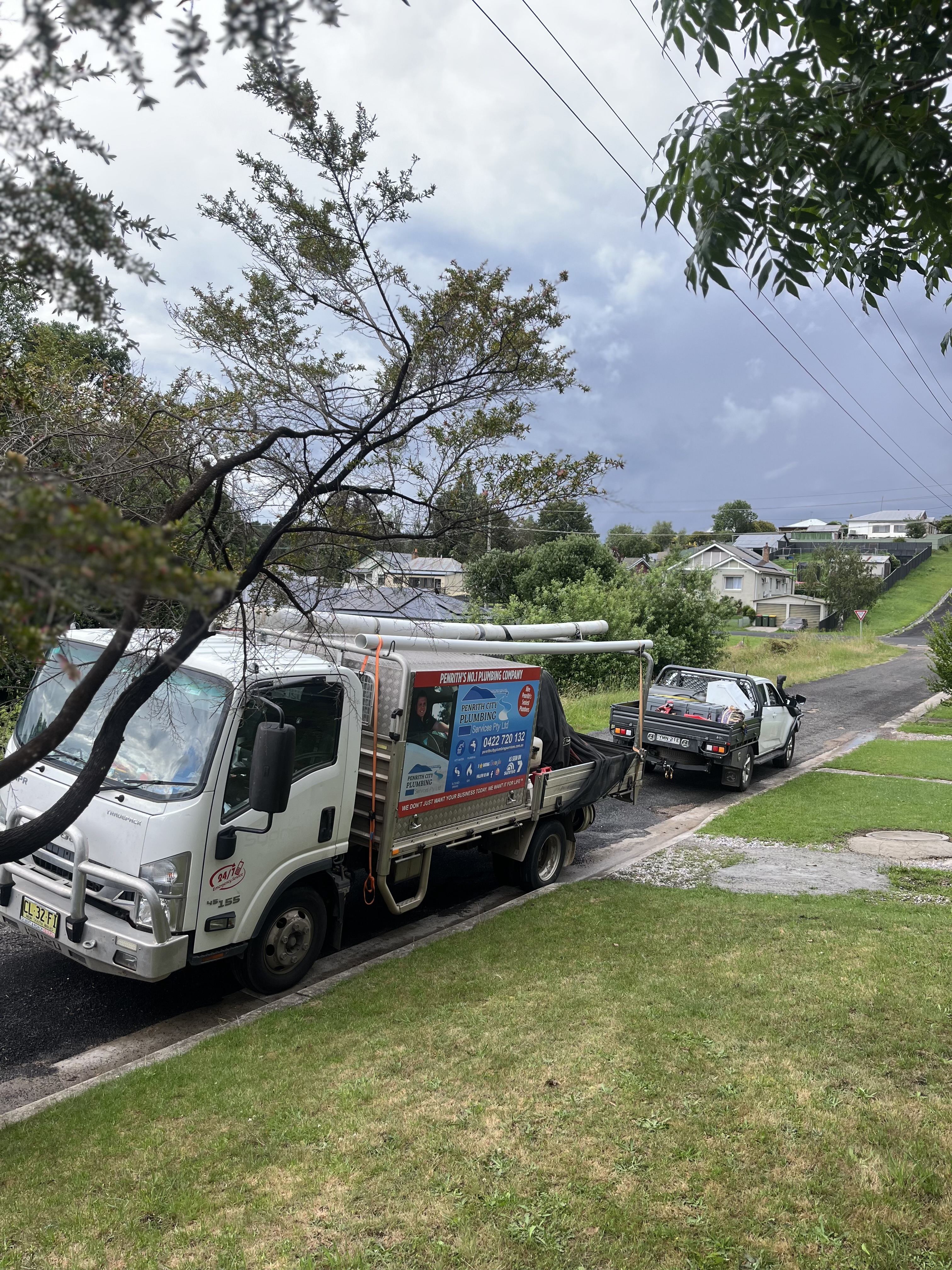 The image shows a service vehicle from Drain Clearing Penrith parked on a residential street in Mulgoa. The truck appears to be well-equipped for plumbing services, possibly indicating readiness for drain clearing. The surroundings feature a well-kept lawn and tree, with houses visible in the background, suggesting a suburban environment. The cloudy sky might indicate a change in weather, but the area appears peaceful. This setting aligns well with the service area of Mulgoa and Penrith, showcasing the company's accessibility for local plumbing needs.