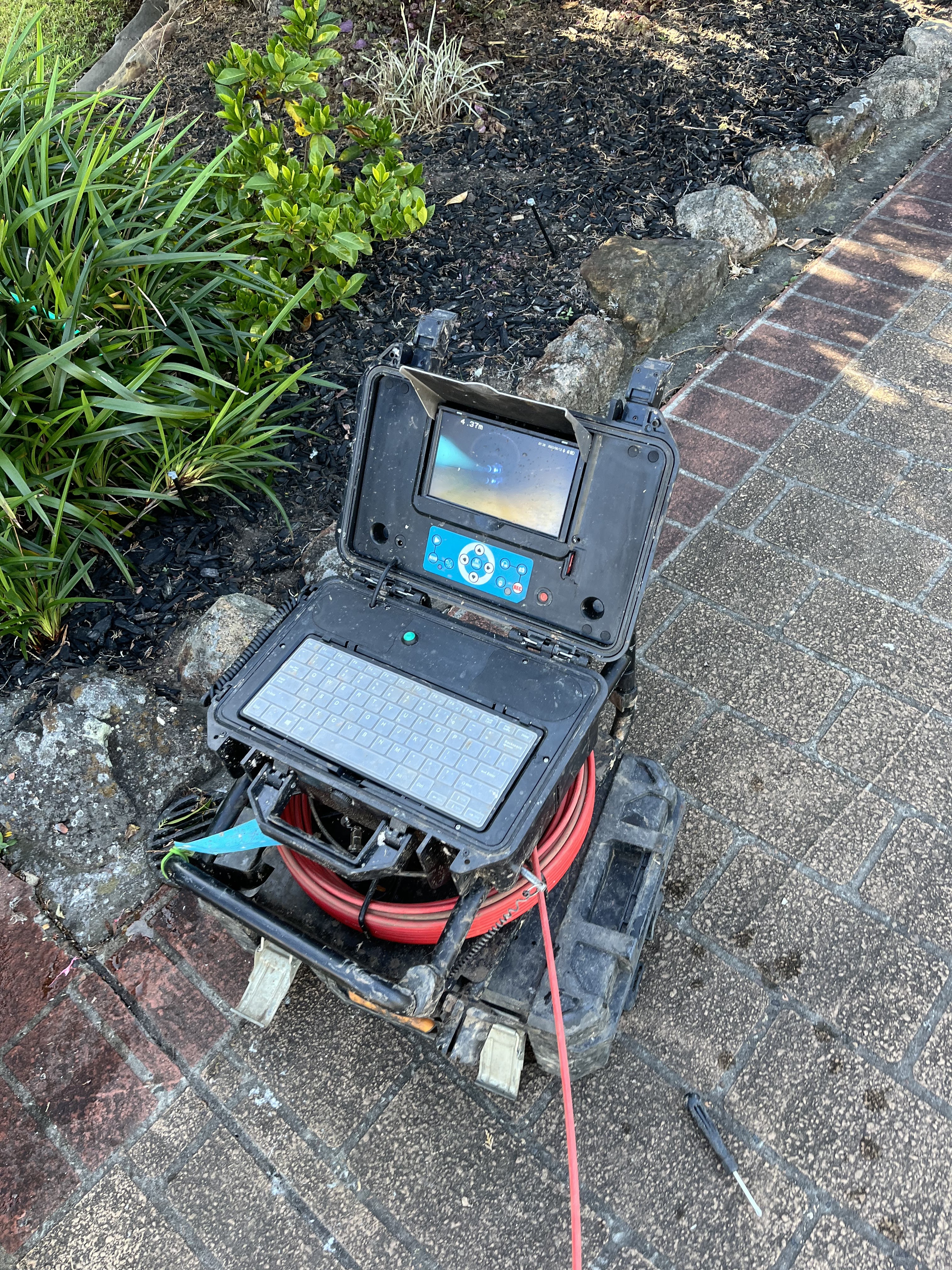 The image features a high-tech drainage inspection camera set up on a sturdy carrying frame. The inspection device, which includes a small monitor and a keypad, indicates it is designed for exploring plumbing systems. The red cable suggests this is a flexible camera that can navigate through pipes to identify blockages or other issues. The background is landscaped with green plants and stones, indicating an outdoor setting likely related to plumbing work.