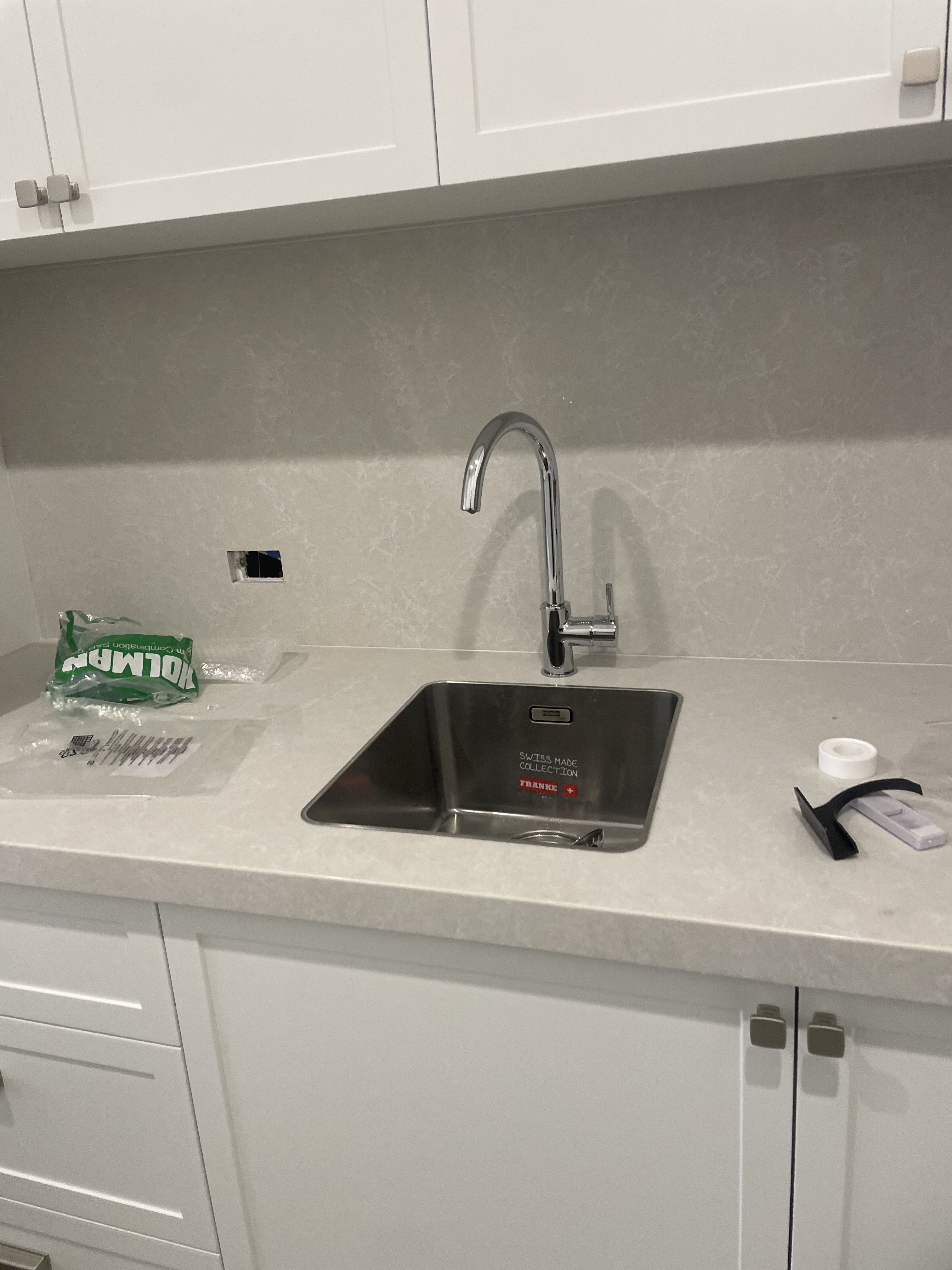 The image features a modern kitchen sink area that appears newly installed or in the process of being finalised. The focus is on a stainless steel sink, which is positioned under a sleek, single-handle tap. The surrounding countertops are made from a light-coloured stone material, complementing the white cabinetry above and below. There is an unpackaged bag of plumbing fittings or accessories on the countertop alongside the sink, indicating that work is ongoing. The wall behind the sink is smooth and clean, hinting at a recently renovated space. This setting is typically relevant for Penrith City Plumbing Services, where they may have just completed plumbing installation or renovation work for a kitchen in Mulgoa, emphasising their expertise in delivering quality plumbing solutions in residential areas.