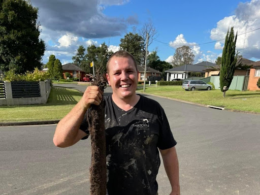 In the image, a cheerful plumber is standing on a residential street, holding up a large bundle of tangled hair and debris, likely removed from a drain or sewage line. He is dressed in a dark shirt and appears to be covered in some dirt, suggesting he has just finished a tough job. The background shows a well-manicured lawn and suburban homes typical of the Mulgoa area in New South Wales. This scene highlights the successful completion of a plumbing task, showcasing the plumber's role in maintaining clean and functional drainage systems for residents. His smile indicates satisfaction with his work, reflecting the customer service focus of Penrith City Plumbing Services, which prides itself on reliability and professionalism in serving the local community.