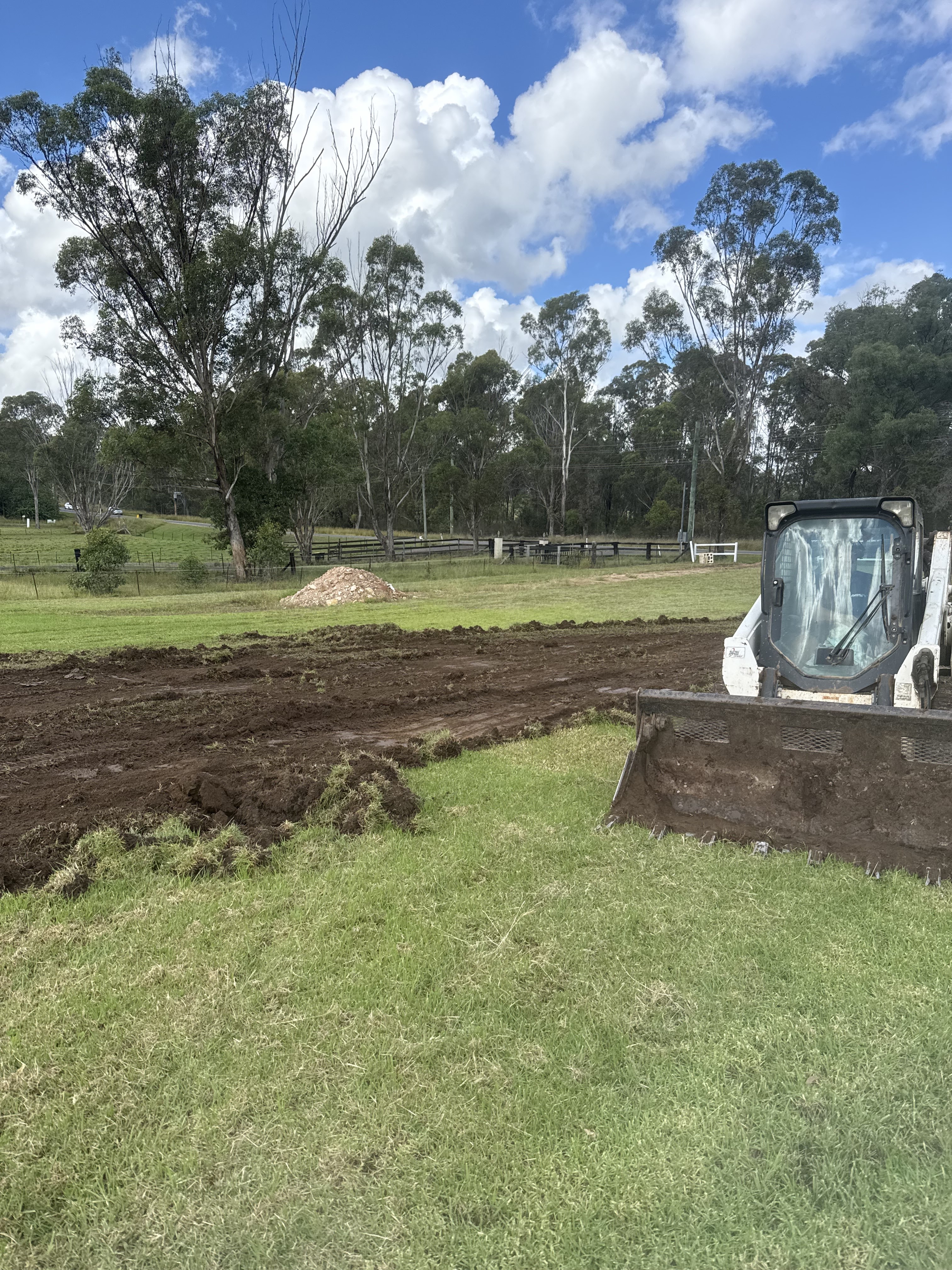The image shows a construction site with a small bulldozer positioned in the foreground. It's operating on a grassy field, indicating that land preparation is underway, possibly for plumbing infrastructure installation. In the background, there are several trees and an open area, suggesting a rural or semi-rural setting in Mulgoa, NSW. The earth appears freshly turned over, which could indicate preparation for utility lines or drainage needs, aligning with Penrith City Plumbing Services' expertise in plumbing and drainage solutions. This imagery implies a site ready for plumbing works, reinforcing the business's commitment to providing efficient plumbing services in the area.