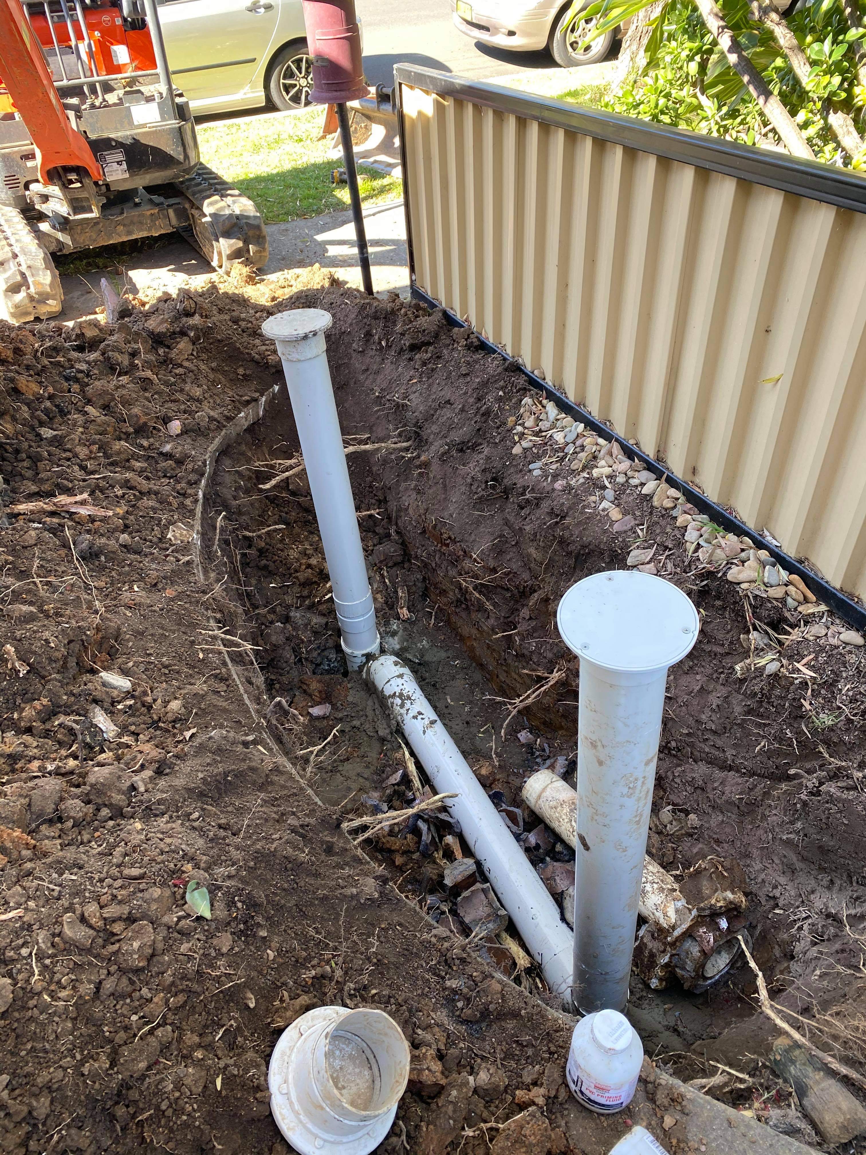 The image depicts a plumbing installation site where two grey drainage pipes are positioned upright in freshly excavated earth. The soil around the pipes is dark and loose, suggesting recent digging. The pipes appear newly installed, possibly intended for drainage or sewer connections. In the background, some machinery is partially visible, likely a mini excavator used to facilitate the trenching work. This scene indicates active plumbing work, fitting within the services that Penrith City Plumbing Services offers. Ensuring proper drainage systems is essential for residential and commercial properties, and this installation suggests the company is addressing plumbing needs in Mulgoa, NSW, or surrounding areas.