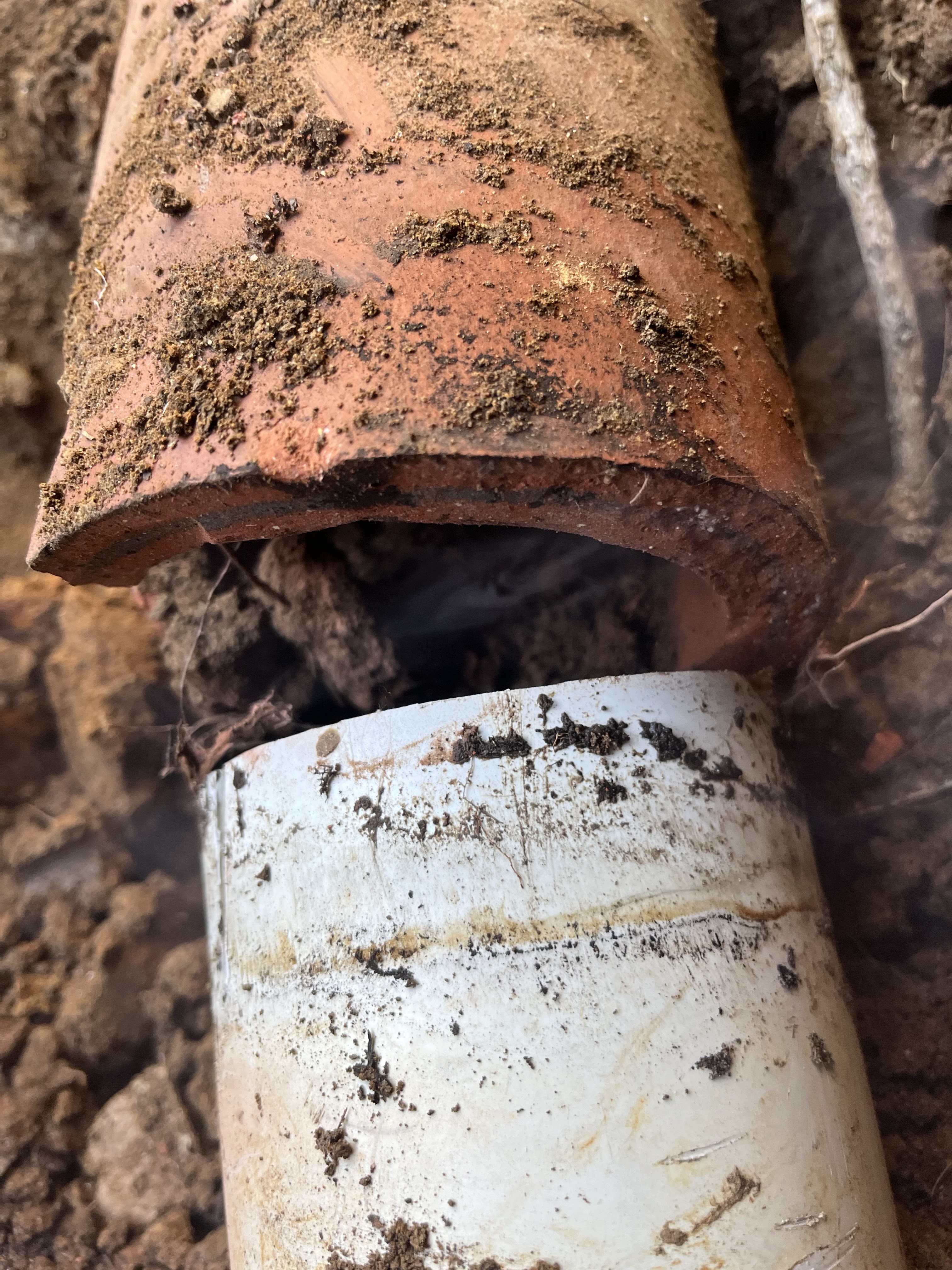 The image depicts a section of plumbing, showcasing a clear issue involving a transition between two types of pipes. On the left, there is a terracotta pipe, which is showing signs of decay and may be contributing to blockage or leakage issues. On the right, there is a newer white PVC pipe, typically used in modern plumbing systems. The two pipes are partially connected, indicating an area where plumbing work may be required to properly join or replace the older terracotta section. The surrounding soil suggests that this plumbing is buried underground, possibly in a residential area serviced by Penrith City Plumbing Services. This kind of work is essential for preventing water leaks or backup in drainage systems, highlighting the importance of professional plumbing services in maintaining efficient and reliable plumbing systems in Mulgoa and surrounding areas.