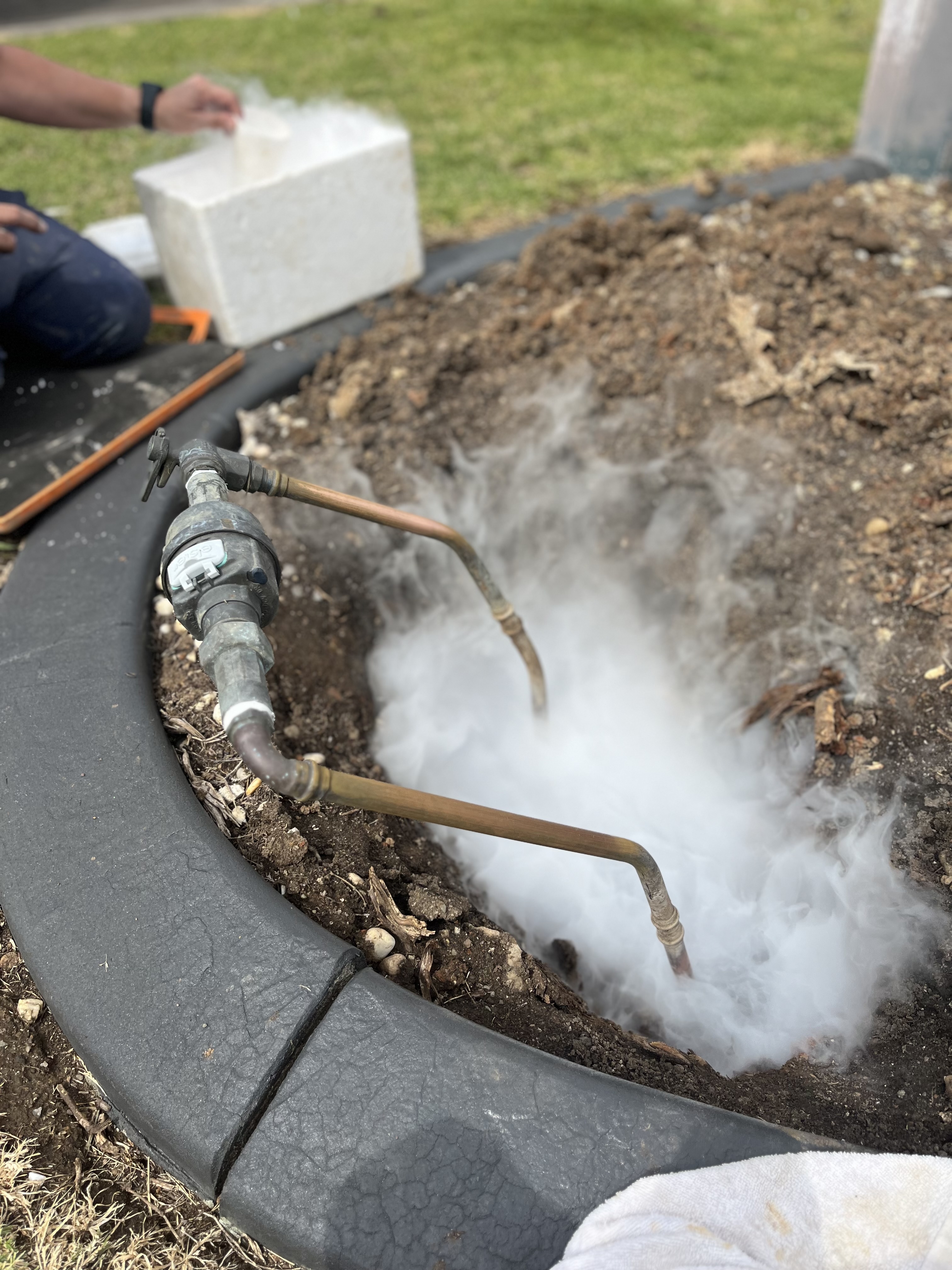 The image depicts a plumbing service in action, focusing on a section of ground that appears to be disturbed, likely indicating excavation. Visible are copper pipes emerging from the ground, which are connected to a plumbing fixture. There is a significant amount of steam or smoke billowing from the area, suggesting that heat or pressurised air is being released—this could indicate testing for leaks or preparing the site for further plumbing work. In the background, a technician is seen working with equipment or tools, likely meant to assist in the plumbing task at hand. This scenario typically involves diagnostic work or treatment of the plumbing system, ensuring that services provided by Penrith City Plumbing are efficient and effective for their clients in Mulgoa and the surrounding areas.