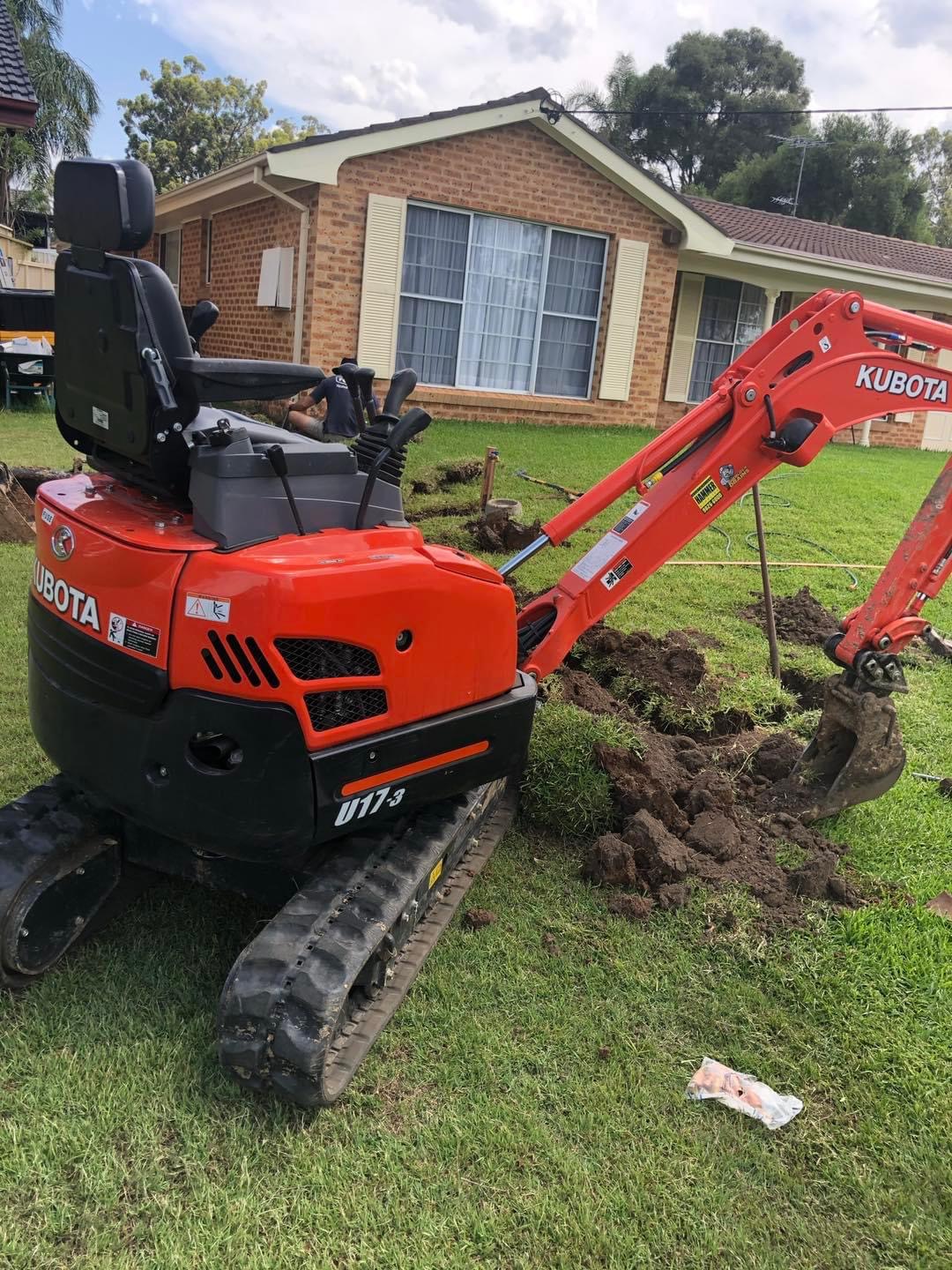 The image shows a compact Kubota excavator, specifically the U17-3 model, positioned on a neatly maintained patch of grass outside a residential home. The excavator is actively digging, likely for plumbing installation or repair work, which could include laying new pipes, fixing drainage issues, or preparing a site for an outdoor plumbing feature. The operator appears to be focused on the task at hand, indicating that this could be part of a plumbing service by Penrith City Plumbing Services. The house in the background suggests a suburban setting, typical of the residential areas served by the business in Mulgoa and surrounding locations.