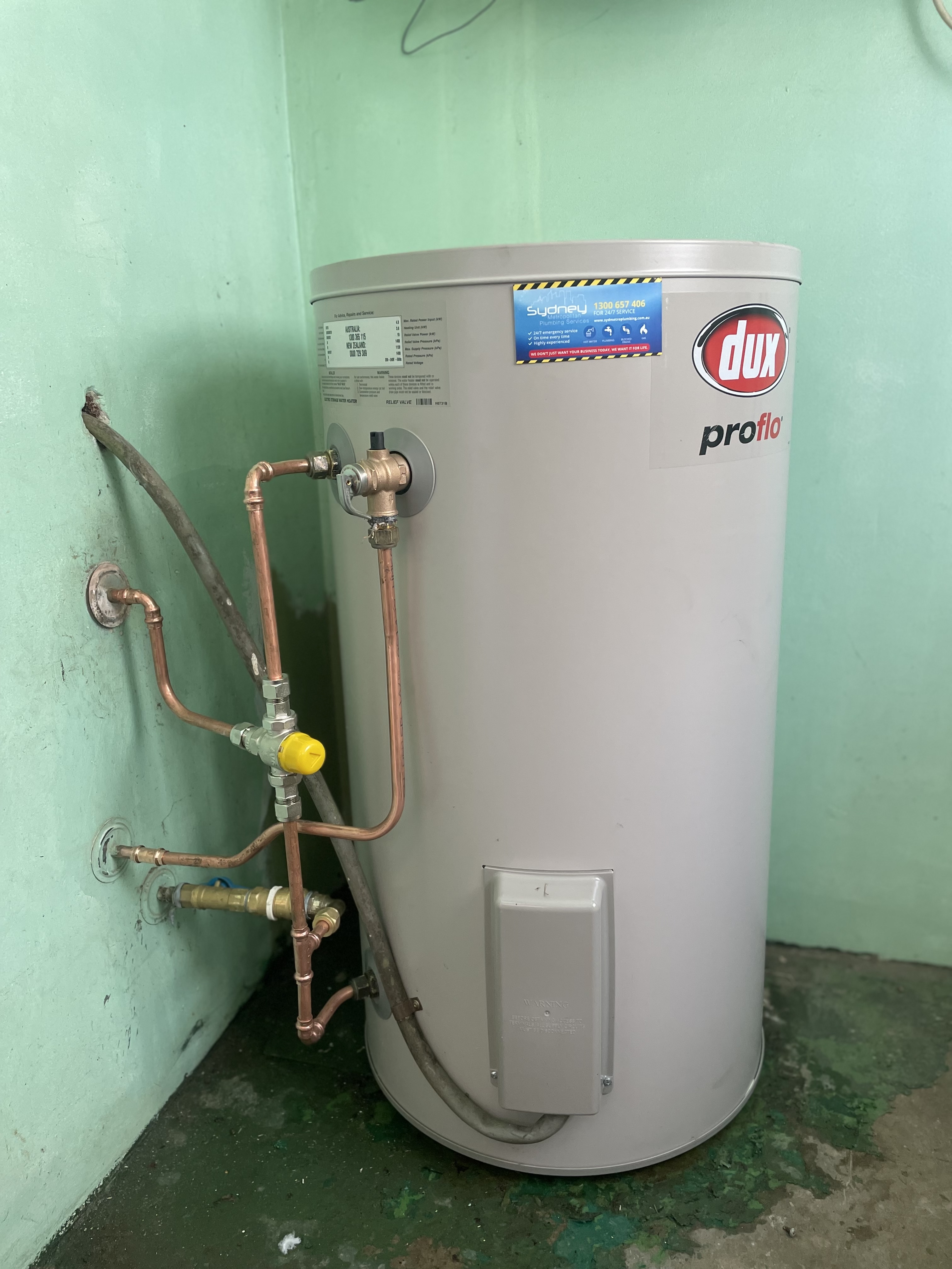 The image features a cylindrical hot water system, specifically a Dux Proflo model, positioned against a green wall. The unit is connected to pipes that supply hot and cold water, demonstrating its integral role in home plumbing. There are copper pipes that are attached to the water heater, facilitating the flow of water into and out of the system. This installation showcases a typical plumbing fixture that Penrith City Plumbing Services would install or service, highlighting the importance of reliable hot water systems in residential settings, particularly in Mulgoa, NSW. The clean installation suggests a professional job, potentially indicative of the quality service offered by the business.