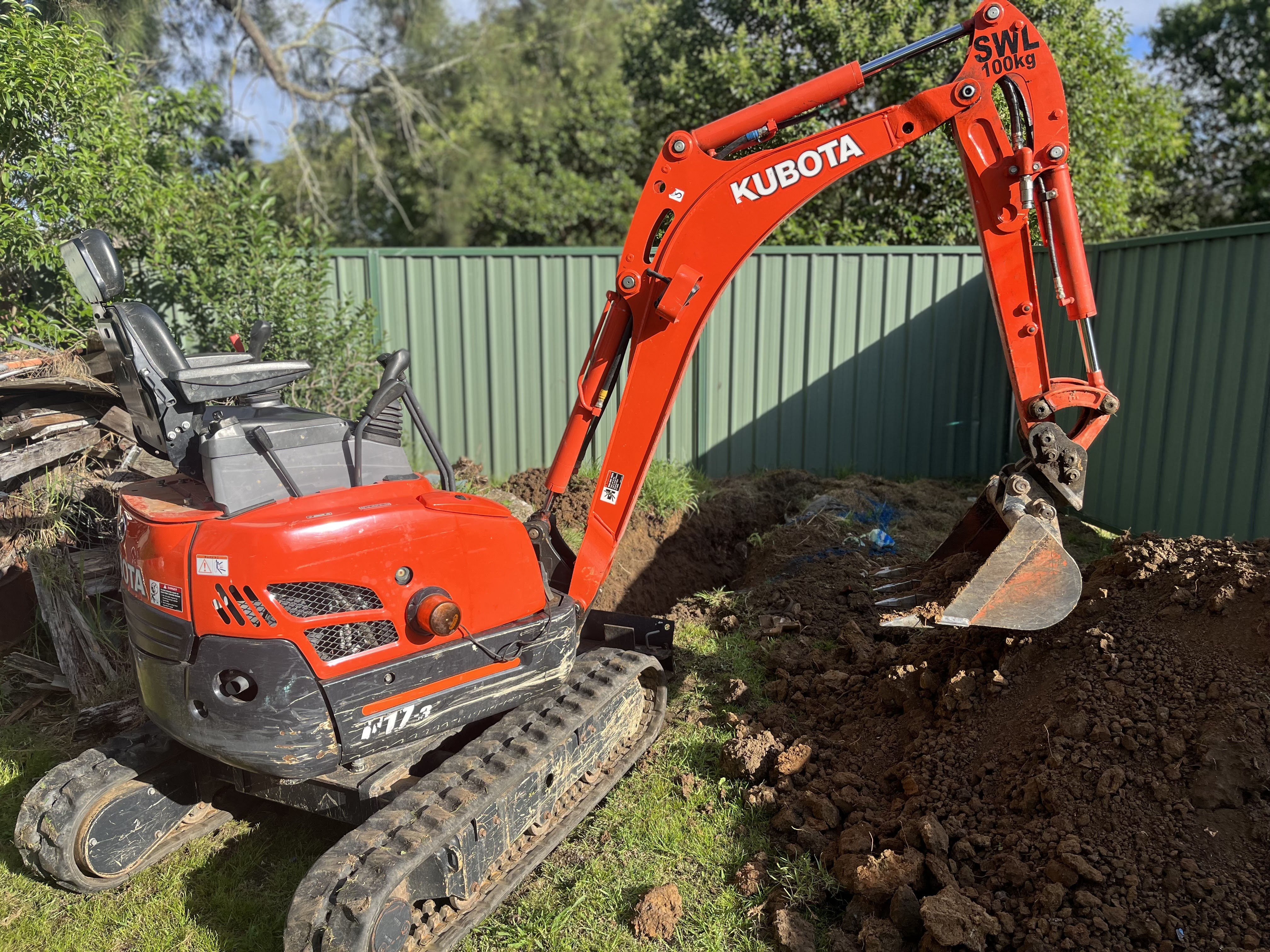 The image depicts a Kubota mini excavator parked in a residential setting, surrounded by a green fence and a mound of freshly dug earth. This machinery is likely being used by Penrith City Plumbing Services for excavation work related to plumbing installations or repairs. The excavator's digging arm is positioned to the side, indicating that recent work has been completed or is in progress. The presence of the excavator suggests that the plumbing services offered include larger scale jobs that require ground excavation, such as installing pipelines or repairing sewer lines, particularly in the Mulgoa area and surrounding regions.