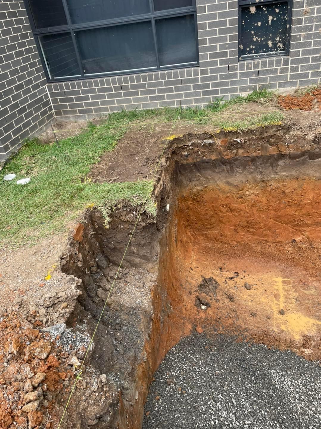 The image displays an excavation site where a trench has been dug revealing distinct layers of soil and substrate. The sides of the trench show a clear demarcation between the topsoil and the deeper earth layers, indicative of preparatory work for plumbing installation or repair. Nearby, the surrounding grass area shows signs of disturbance from the excavation process. It is likely that this site is being prepared for pipeline installation, possibly for sewerage or drainage systems, which is essential for plumbing services provided by Penrith City Plumbing Services. The precise digging suggests careful planning to avoid existing structures and ensure effective plumbing solutions in Mulgoa, NSW.