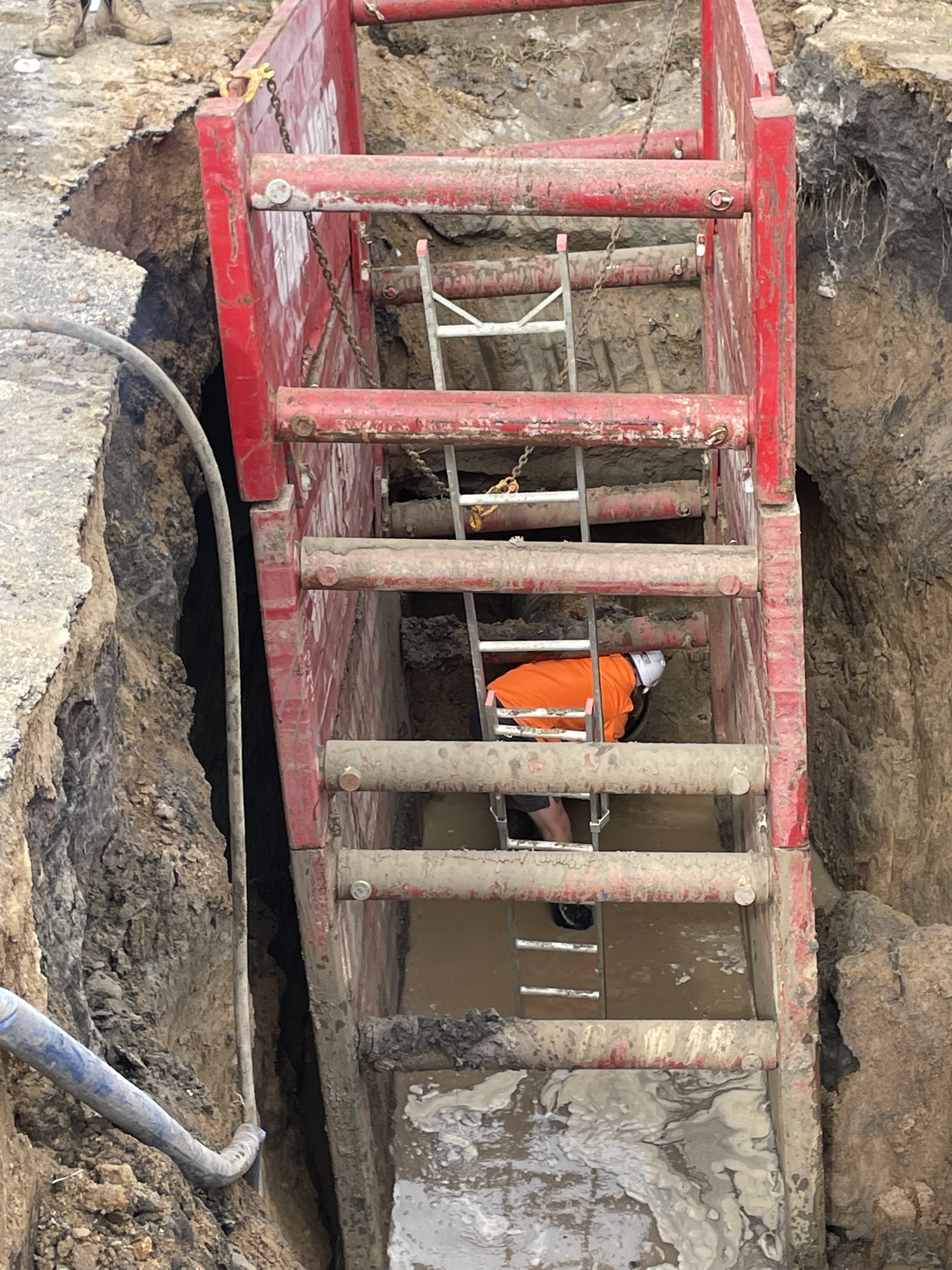 The image shows a construction site where a plumber is working in a deep trench, surrounded by red temporary fencing or shoring. The trench appears to be dug out for plumbing installation or repairs, with a significant amount of water accumulating at the bottom, indicating possible drainage issues or groundwater presence. The plumber, dressed in safety gear, including an orange shirt, is positioned inside the trench, where they are performing tasks to resolve plumbing problems. The site looks to be securely managed, with the use of a ladder for safe access and egress. This setting is typical for the Penrith City Plumbing Services, reflecting their commitment to ensuring thorough and safe plumbing installations for residents in areas such as Mulgoa and surrounding suburbs.