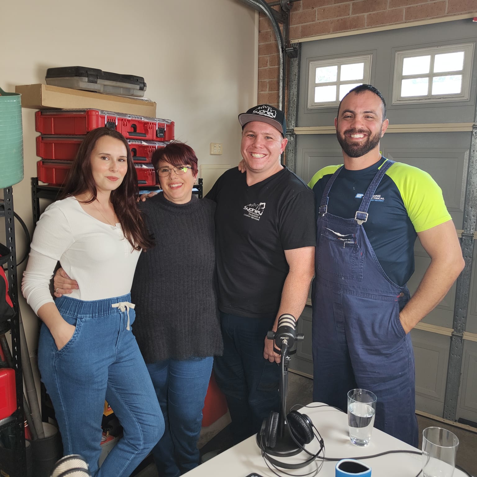 The image features four individuals posing together in a garage-like setting filled with tools and equipment that suggests a work environment related to plumbing or home maintenance. The two women and two men are smiling, conveying a friendly and approachable atmosphere, likely indicative of a team from Penrith City Plumbing Services. The garage backdrop may include plumbing supplies or tools being prepared for job assignments, potentially representing their readiness to tackle plumbing challenges. This visual showcases the team spirit and professionalism of the business, highlighting their commitment to service in Mulgoa and surrounding areas.
