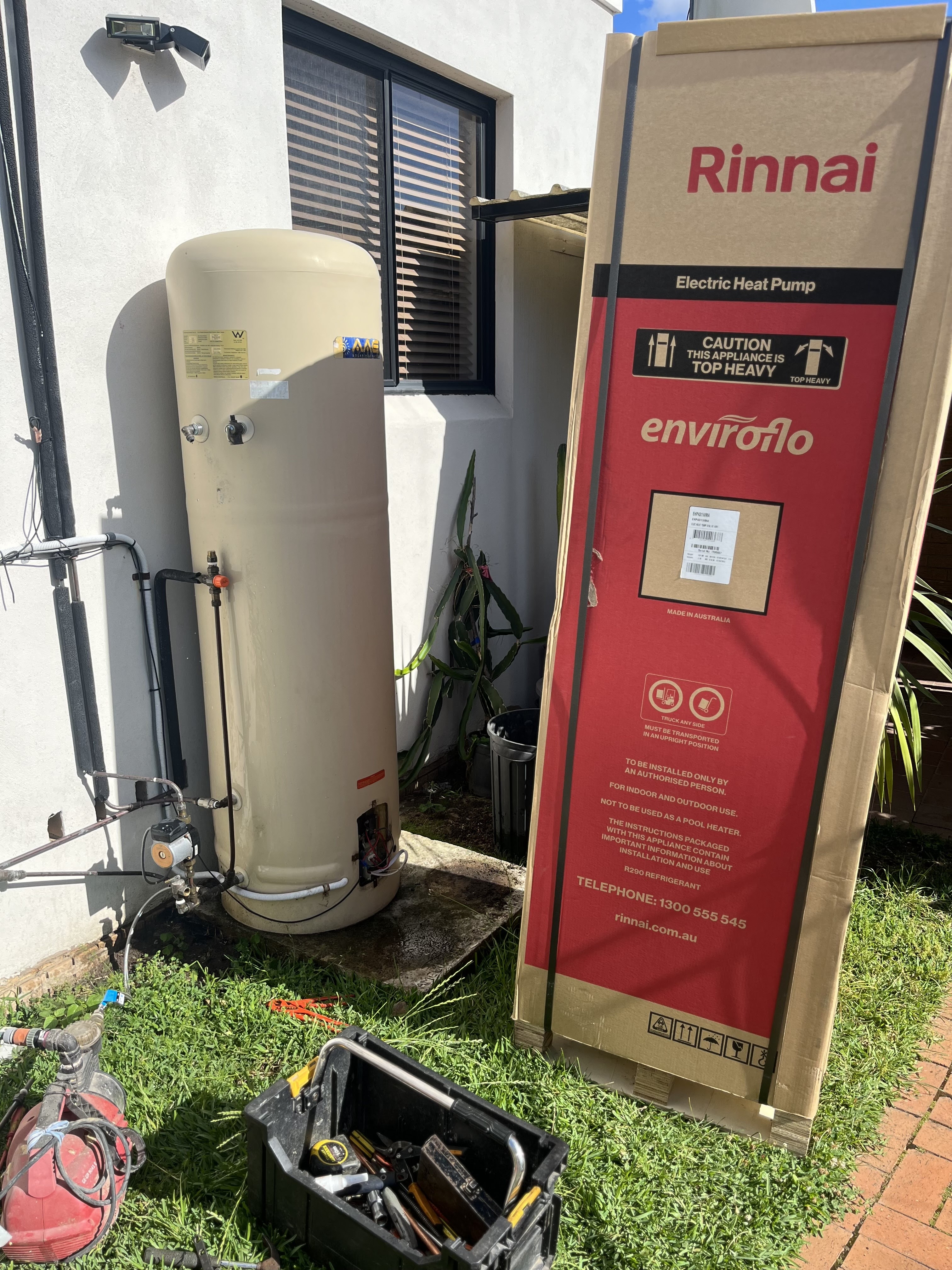The image depicts an outdoor area where a new Rinnai electric heat pump is being installed next to an existing water heater tank. This setting is typical for a plumbing service as it showcases the installation process of a hot water system. The heat pump is in its packaging, indicating that it is either ready for immediate installation or has just arrived at the site. Surrounding the area, tools are visible, likely from the plumbing kit, indicating that a professional plumber from Penrith City Plumbing Services is on-site, preparing to execute the installation. The location likely serves a residential property in Mulgoa, NSW, illustrating the business's commitment to servicing local homes with efficient hot water system solutions. The presence of grass and the positioning of plumbing equipment also highlight the outdoor nature of this plumbing task, a common part of their services.