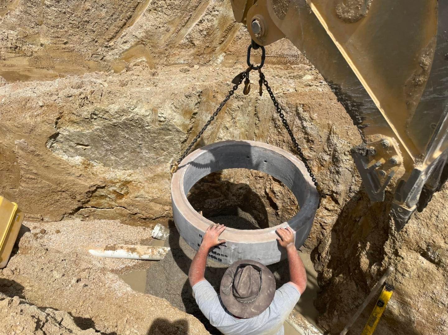 The image features a construction scene where a worker is positioning a large concrete pipe, likely a sewer or stormwater pipe, into a trench. The worker, wearing a grey shirt and a wide-brimmed hat for sun protection, appears to be carefully guiding the pipe as it is hoisted by an excavator's crane. The trench is surrounded by earth and appears to be related to plumbing work, possibly for upgrading or installing a drainage system. This activity is crucial in plumbing services, ensuring proper water flow and infrastructure in the Mulgoa area and surrounding suburbs.