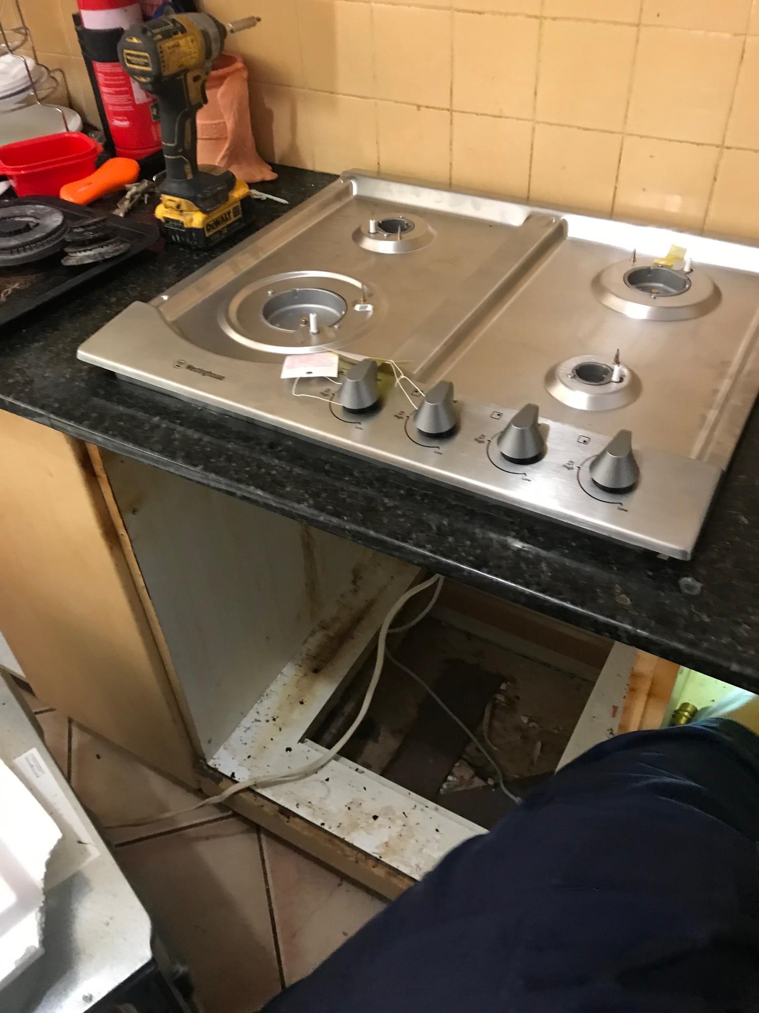 The image shows a kitchen environment with a partially installed stainless steel gas cooktop sitting on a black granite countertop. The area beneath the cooktop appears to be unfinished, revealing exposed wiring and pipes, which indicates either ongoing plumbing or electrical work. Tools and parts are scattered on the countertop, including what looks like a power drill and other plumbing tools, suggesting that a plumbing service is in progress. This scene may indicate that the Penrith City Plumbing Services team is currently providing installation or maintenance services in a customer’s kitchen in Mulgoa, ensuring that all components are safely and effectively connected, likely following the guidelines for gas appliance installation as mandated by local regulations.