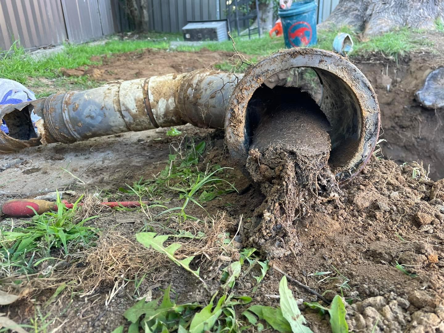The image shows a damaged sewer pipe that has been excavated from the ground. The pipe appears to be heavily corroded and is obstructed by a significant amount of tree roots penetrating through it. This blockage likely reflects poor drainage and could lead to severe plumbing issues, such as sewage backups or leaks that require professional intervention. The visible tools nearby hint at ongoing repair work, demonstrating the need for skilled plumbing services. Penrith City Plumbing Services, located in Mulgoa, NSW, could provide expert assistance in diagnosing and resolving such drainage problems efficiently.
