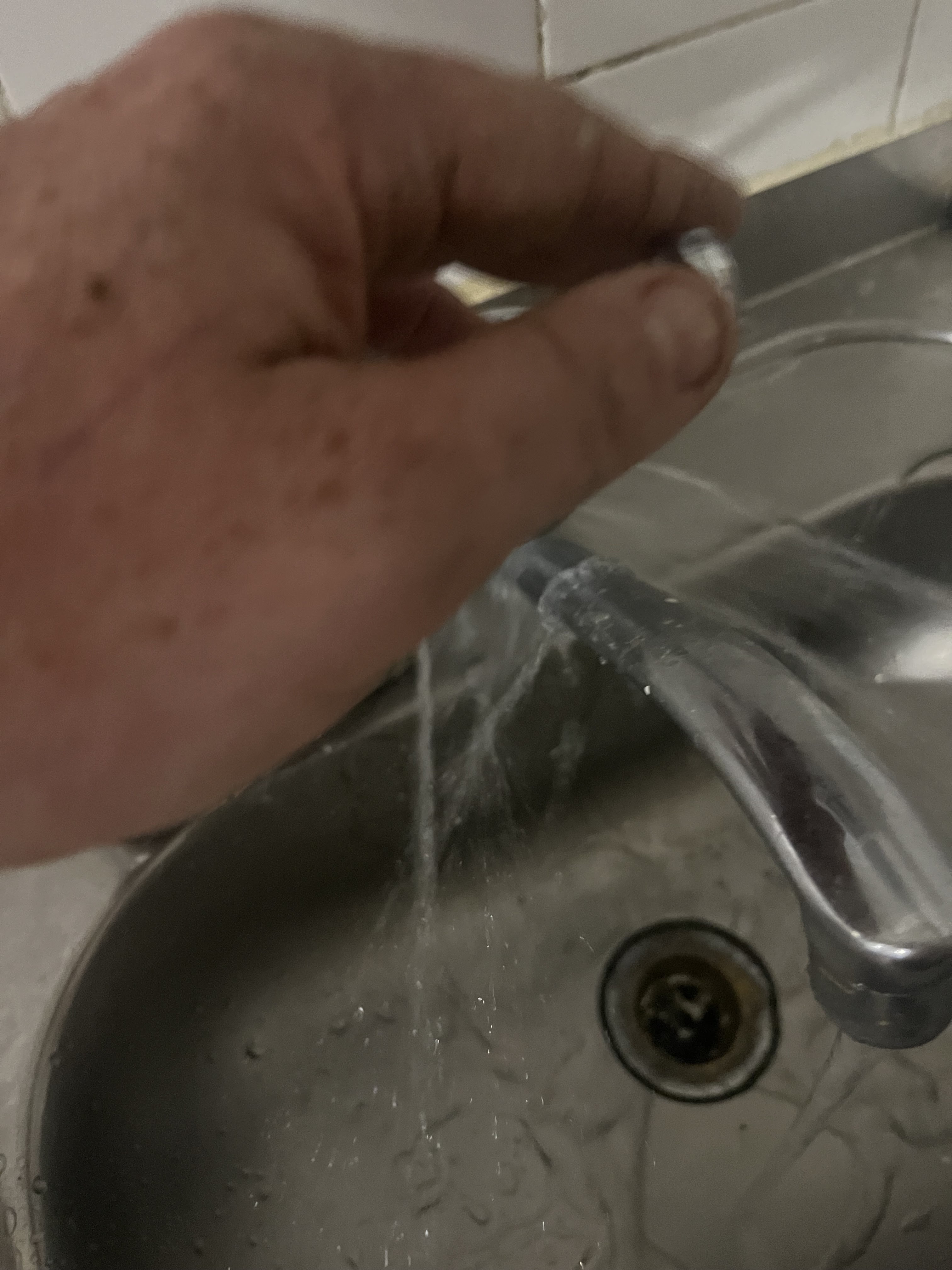 The image depicts a hand reaching towards a faucet in a kitchen sink. Water is actively flowing from the faucet, suggesting that the tap is in use or possibly leaking. The hand appears to be attempting to stop or adjust the flow of water, indicating a potential plumbing issue that may need professional attention. The sink is composed of stainless steel and shows some water droplets around the drain, which may suggest a recent repair or ongoing plumbing issue. This situation is relevant to Penrith City Plumbing Services, as they can address tap repairs, leaks, and kitchen plumbing problems in the Mulgoa area and surrounding locations.