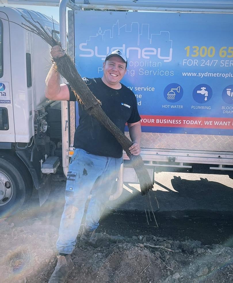 In the image, a cheerful plumber stands proudly beside a service van bearing the logo of Sydney Metropolitan Plumbing Services. He is holding a substantial clump of tree roots, indicating that he has just cleared a blocked drain or sewer line. The surroundings appear to be a work site, possibly in Mulgoa, with soil and debris scattered around, suggesting recent excavation. The bright sunlight creates an uplifting atmosphere, and the plumber's smile reflects satisfaction with a job well done. This scene effectively showcases the hands-on, problem-solving nature of the plumbing trade, highlighting Penrith City Plumbing Services’ expertise in dealing with common plumbing challenges such as root intrusion into drainage systems.