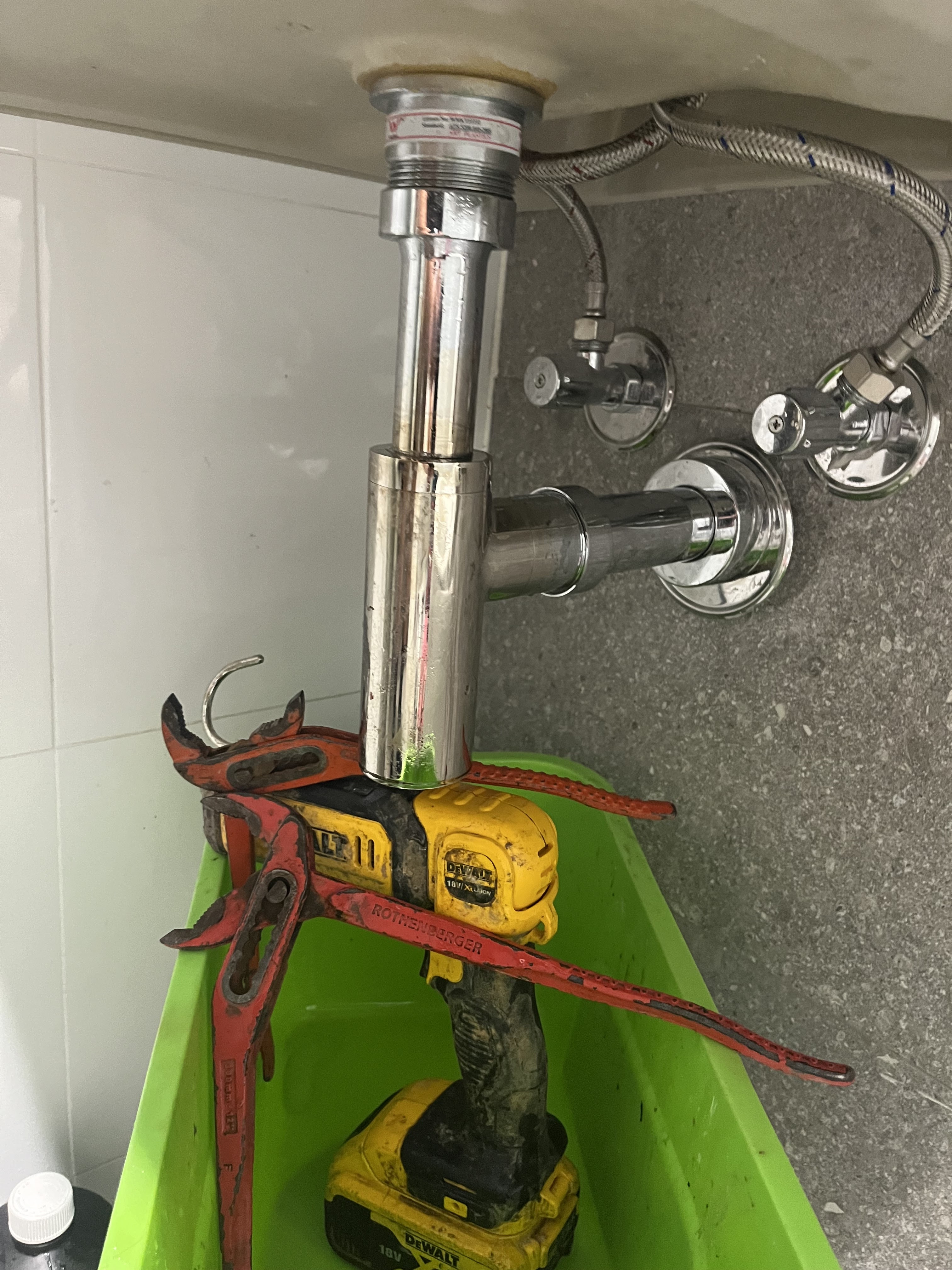 The image displays a plumbing setup beneath a sink, showcasing the components involved in drainage and water supply. In the centre, a shiny chrome-plated pipe connects the sink to the drain, ensuring efficient waste removal. Attached to this is a series of plumbing fixtures, likely for both hot and cold water supply, prominently featuring two shiny chrome valves on the wall, which are essential for controlling water flow. Subtly placed beneath the assembly is a green bucket, probably used to catch any residual water or leaks during the plumbing work. A red pair of adjustable pliers rests on the top pipe, suggesting active maintenance or installation is underway to secure or adjust the fittings. Notably, a cordless drill lies within the bucket, indicating that tasks such as tightening or assembling fittings possibly involved drilling holes or mounting additional components. Overall, this scene reflects the everyday workings of the Penrith City Plumbing Services, highlighting their expertise in handling plumbing installations and repairs in residential settings.