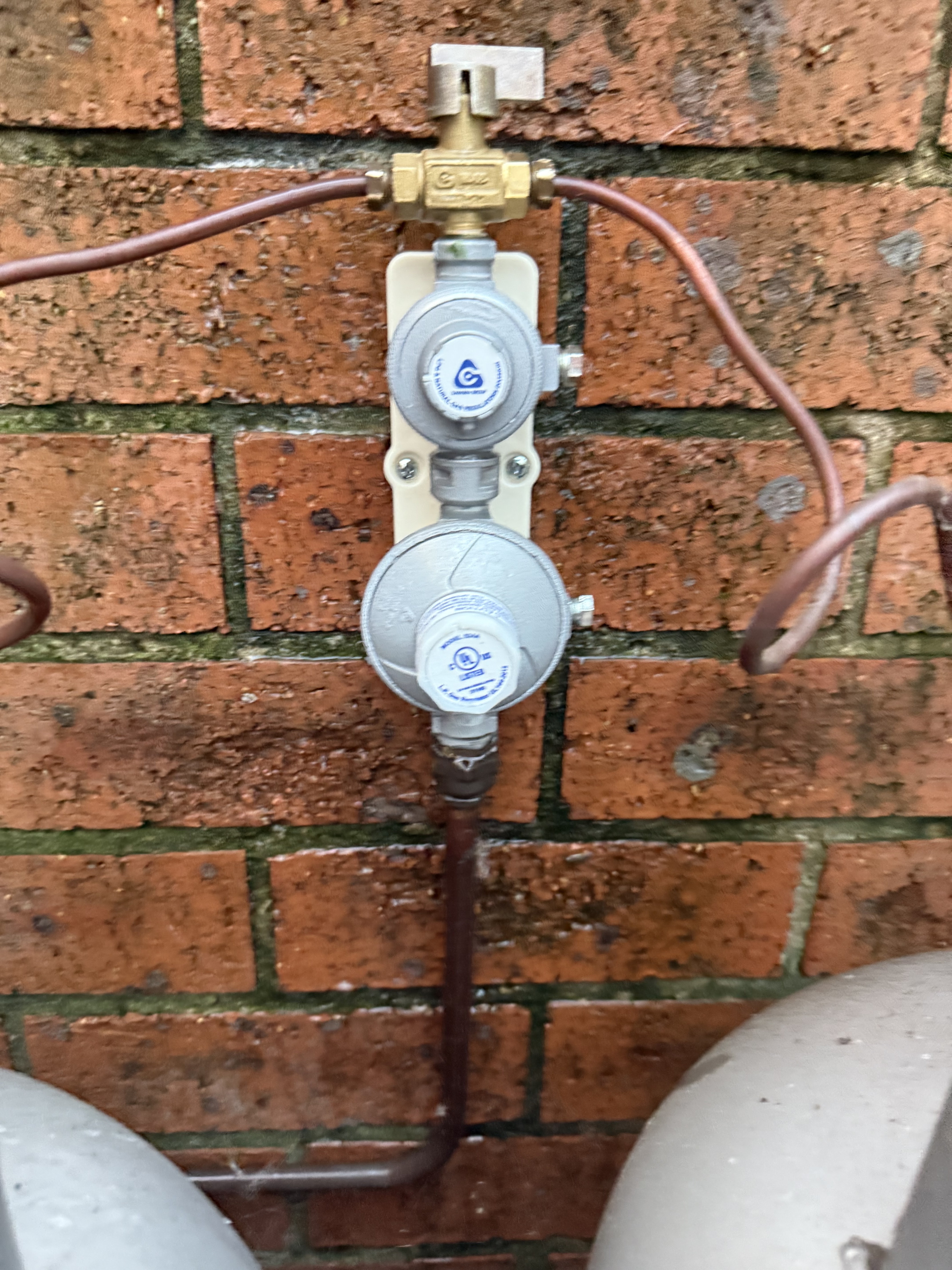 The image shows a gas regulator assembly mounted against a brick wall. The assembly features a brass valve on top, which is used to control the gas flow. Below it, there are two round gas pressure regulators, each with a label that likely specifies the pressure settings for safety. The copper tubing leading downwards connects to the gas lines, which are essential for plumbing systems that involve gas appliances. This image is particularly relevant for Penrith City Plumbing Services, as it highlights aspects of gas plumbing installation and maintenance, which is a critical service they may offer in the Mulgoa area.