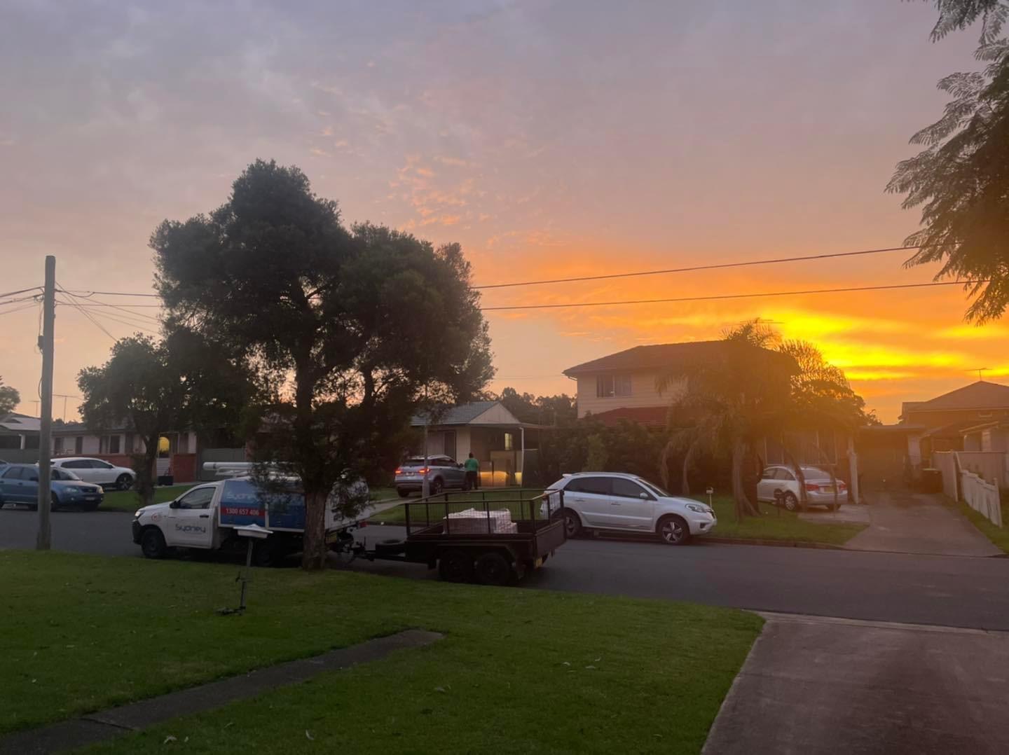 The image depicts a tranquil suburban street during sunset, showcasing a residential area with well-maintained lawns and homes. In the foreground, a plumbing service vehicle is parked next to a trailer, suggesting it is either attending to a job or on a break. The environment conveys a sense of community in Mulgoa, where Penrith City Plumbing Services operates. The warm hues of the sunset could represent the dedication and hard work that the plumbing service provides to local residents, ensuring their plumbing needs are met efficiently before the day ends. The vehicle indicates readiness for emergency services or planned maintenance, reflecting the operational nature of the business.