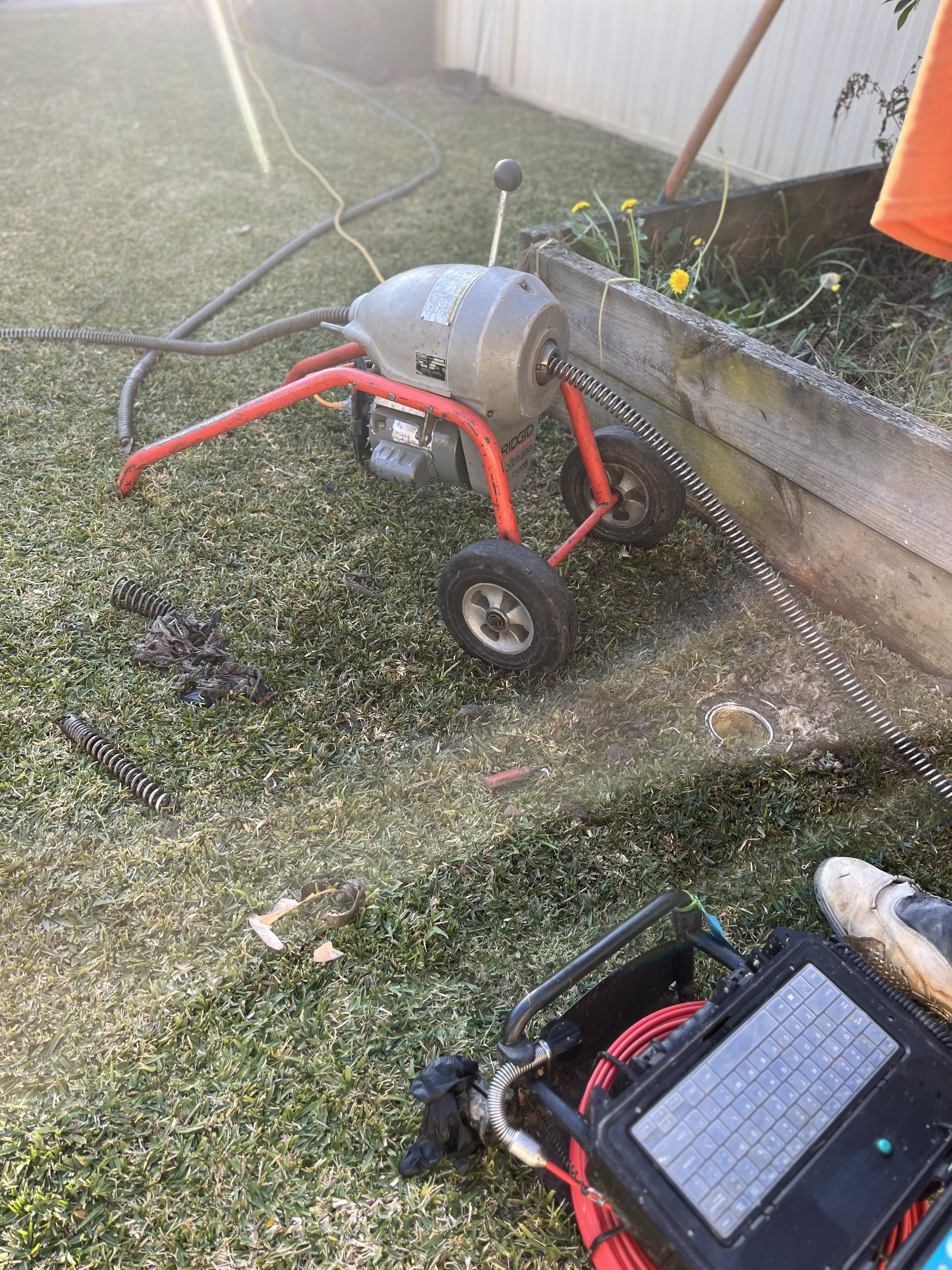 The image features a plumbing tool, likely a drain cleaning machine, set up on a grassy area. The machine appears to be compact and equipped with a small engine, suggesting it's designed for tasks such as unclogging drains. There are coils of plumbing springs, tools, and possibly parts scattered around, indicating an ongoing plumbing job. The background shows a garden area with tools arranged for efficient access, typical of a plumber's work site. The presence of weeds and a wooden border indicates that this area might require attention along with the plumbing work.