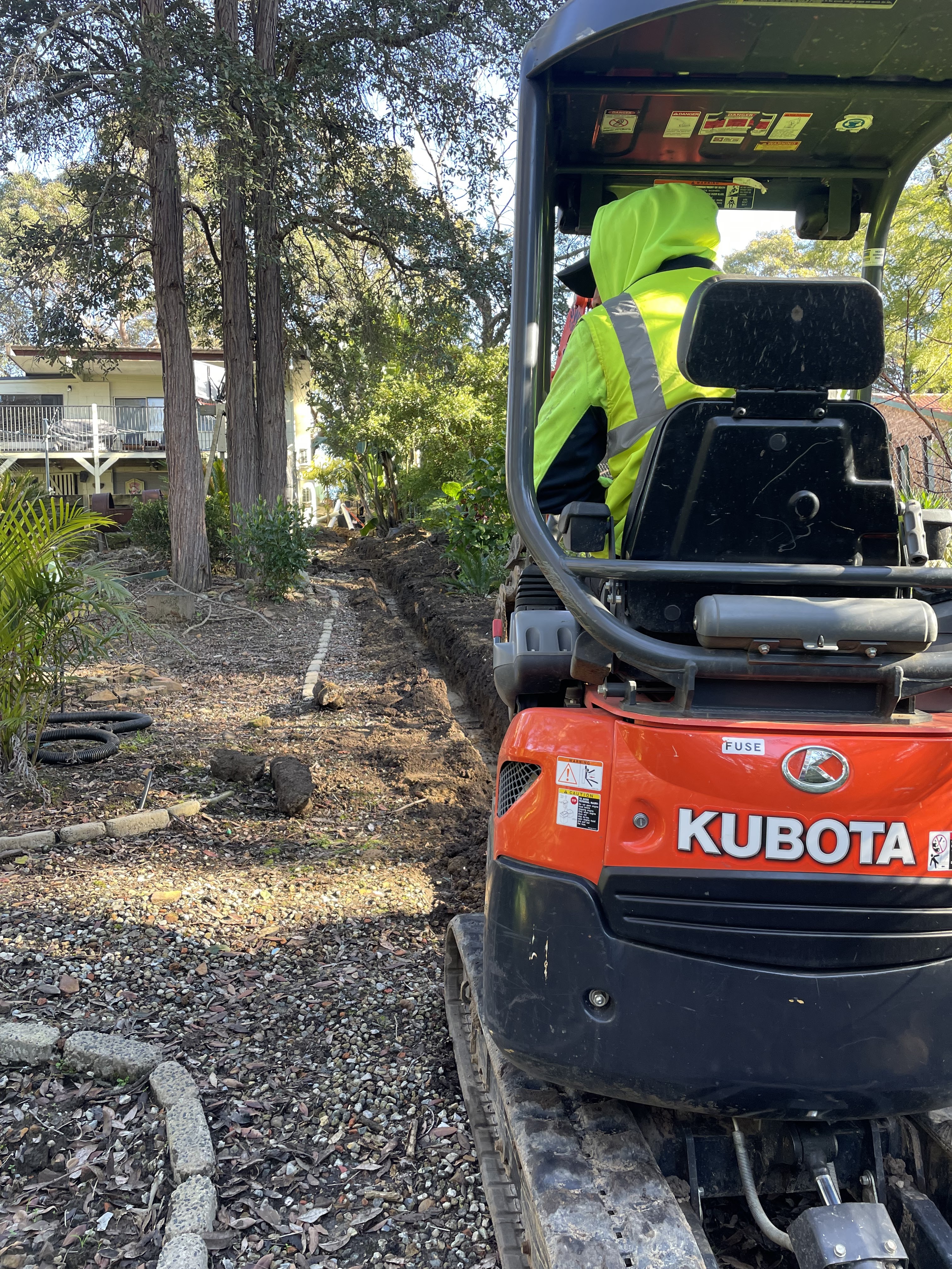 In the image, a worker is operating a Kubota mini-excavator on a residential property. The machine is positioned on a gravel surface, likely to prepare for plumbing installation or repair work. Behind the excavator, a trench has been dug, which may be intended for laying pipes or fixing drainage systems. The area around the excavator is surrounded by overgrown vegetation, suggesting ongoing landscaping or property maintenance. The background features a house, indicating that the job is taking place in a suburban setting, typical for a plumbing service operating in Mulgoa, NSW.
