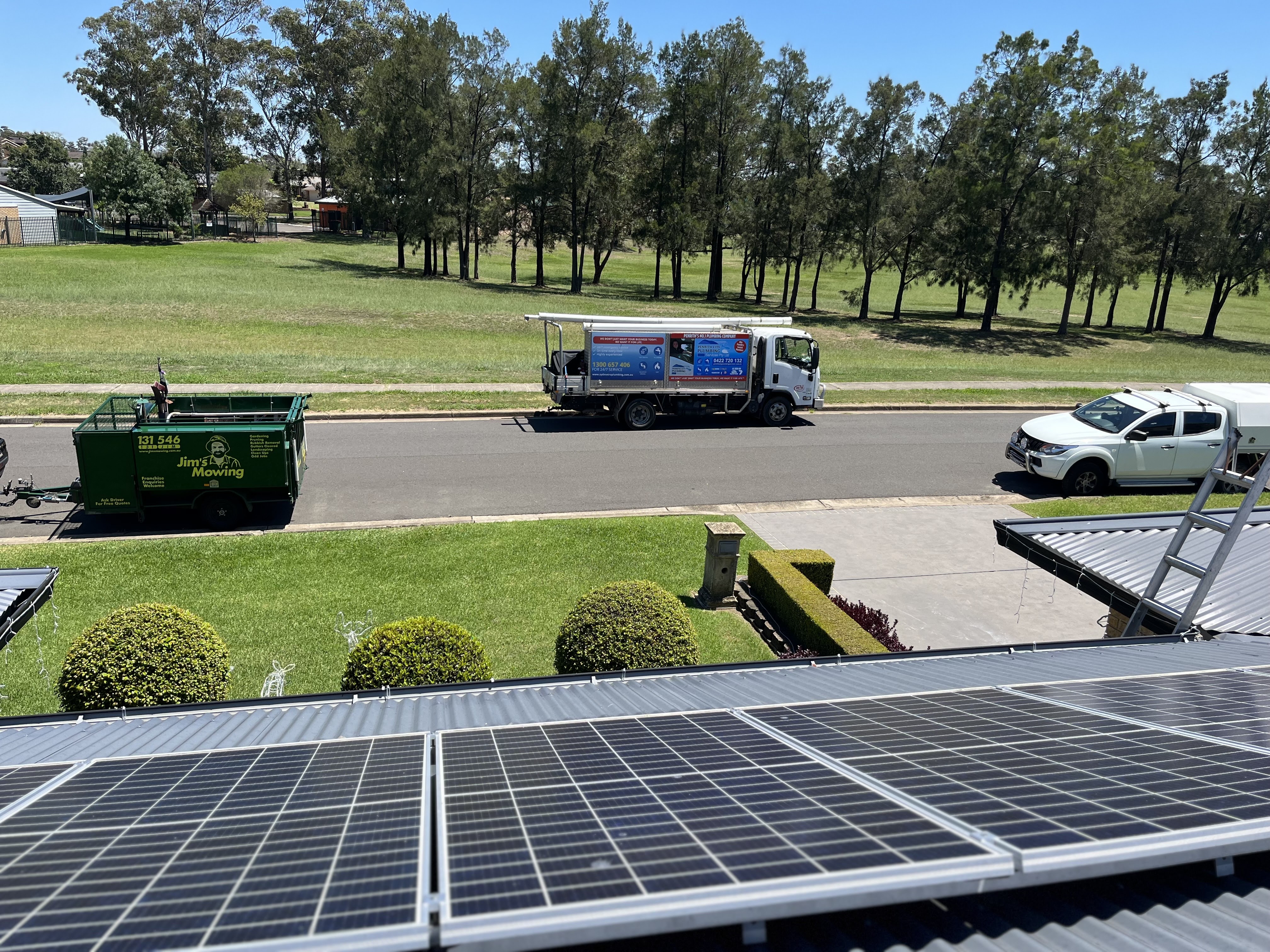 The image depicts a view from the roof of a building with solar panels prominently placed in the foreground. In the background, several vehicles are lined up on a residential street. One vehicle, a truck, appears to be a service vehicle for plumbing or utility services, adorned with advertisements related to plumbing tasks. This suggests that plumbing services, possibly related to renewable energy installations like solar panels, may be part of the offerings by Penrith City Plumbing Services. The surrounding green grass and clear blue sky imply a pleasant day, indicating ideal conditions for external plumbing work or maintenance. Overall, this scene captures a working environment for a local plumber, showcasing both plumbing and sustainable energy services.